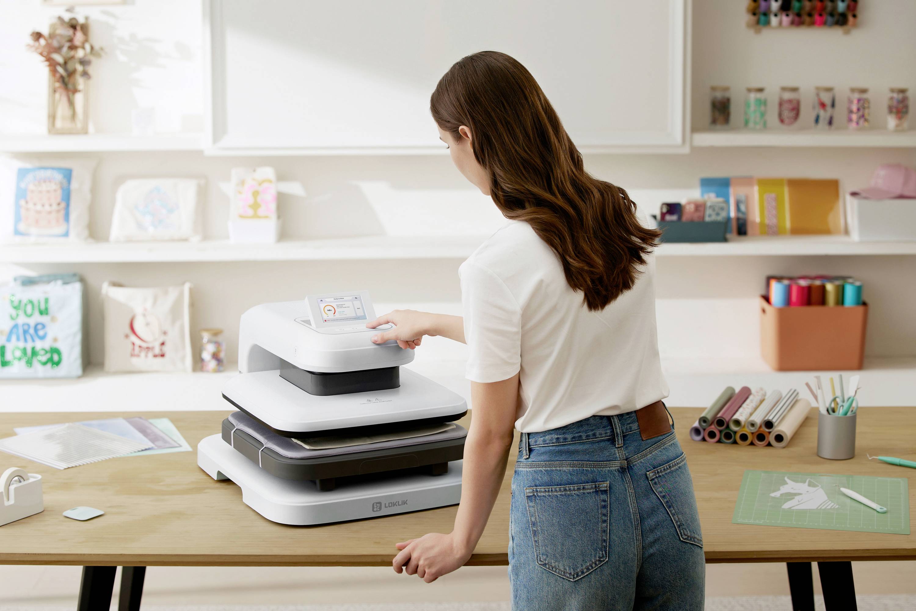 Kobieta stoi przed prasą transferową i obsługuje panel sterowania. Na stole leżą materiały do rękodzieła oraz folie do transferu.