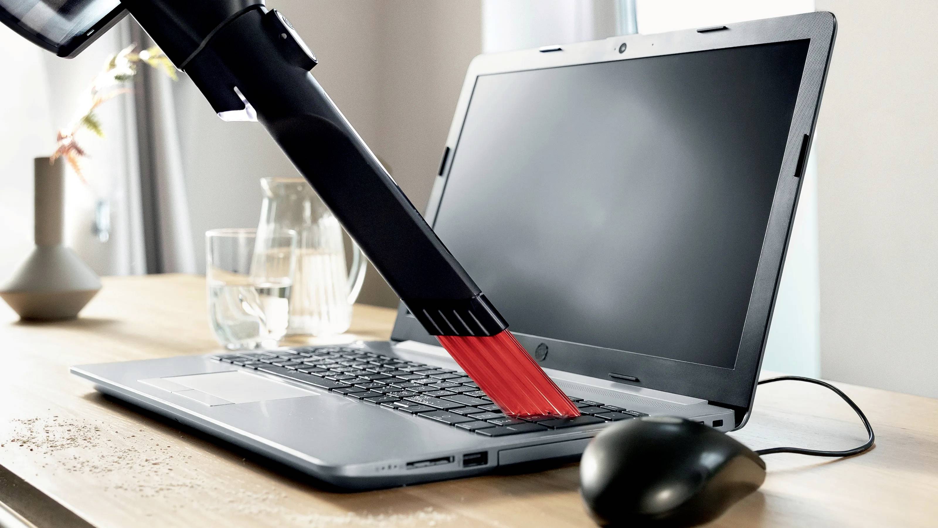 Odkurzacz zbiera okruchy z klawiatury laptopa. W tle widać szklankę wody i oświetlone okno.