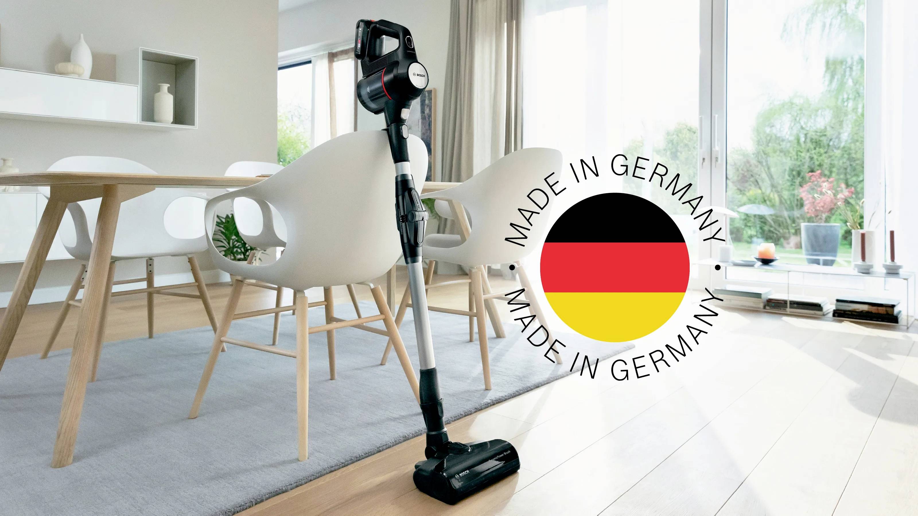 Bezprzewodowy odkurzacz stoi w nowoczesnym, jasnym salonie. Obok znajduje się pieczęć z napisem 'Made in Germany' oraz niemiecką flagą.