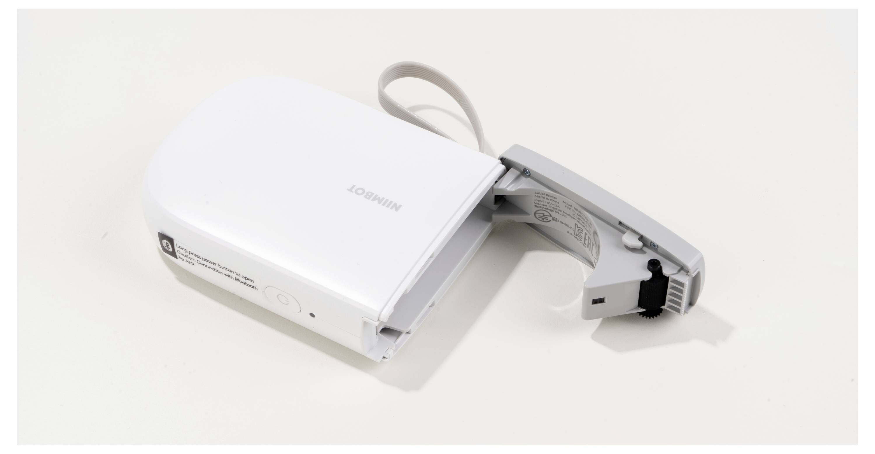 Biały, przenośny sprzęt z uchylną pokrywą i złączem USB, prawdopodobnie drukarka etykiet, leży na białej powierzchni.