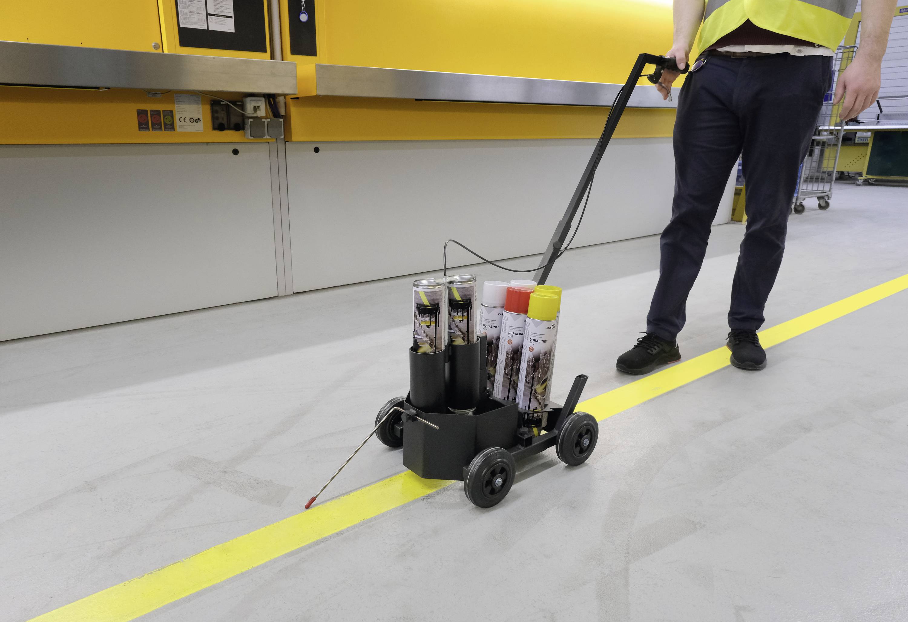 Robotnik ciągnie mały, czarny wózek z puszkami farby przez żółtą linię na podłodze w środowisku przemysłowym.