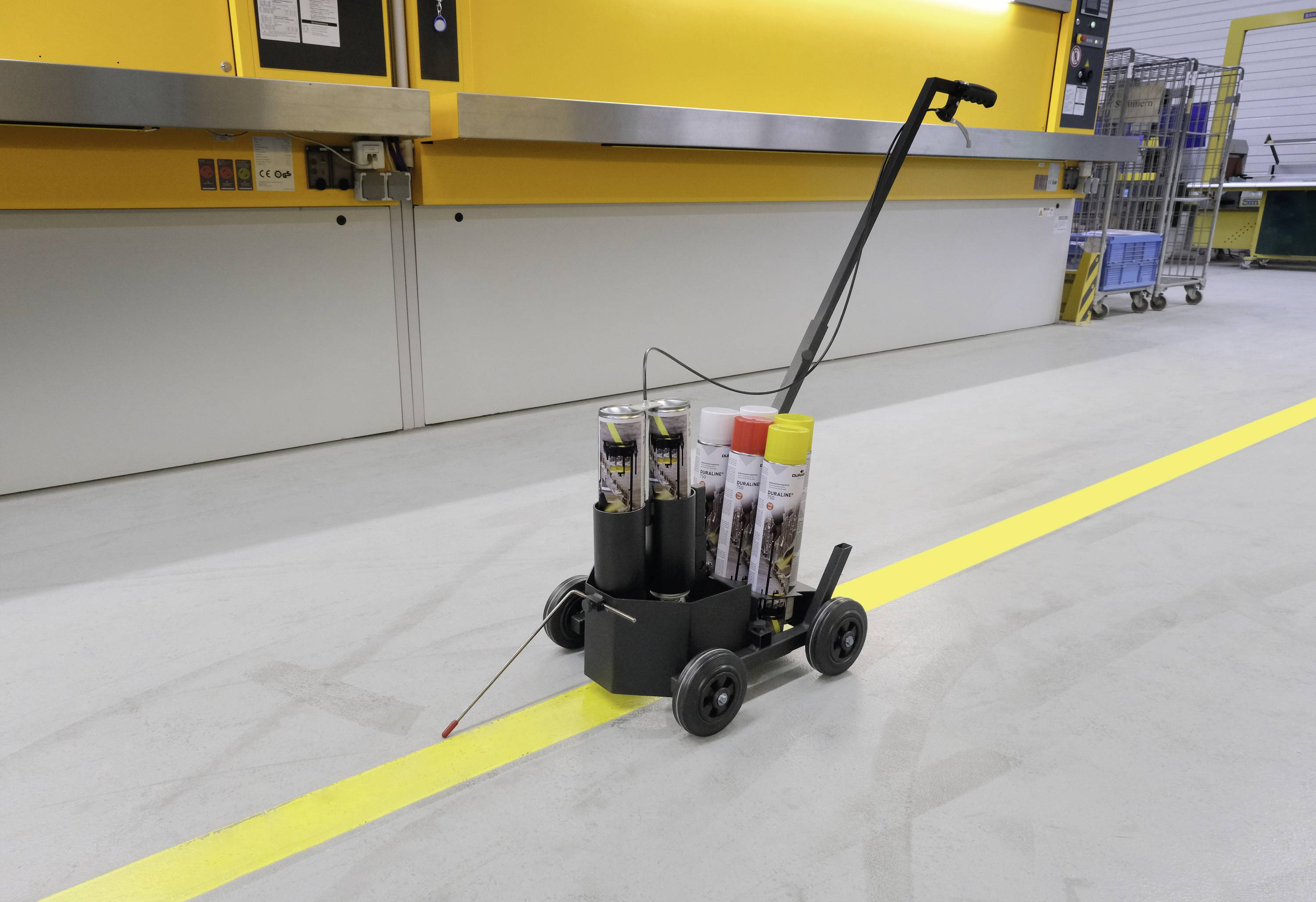 Mały, czarny wózek z uchwytem, przewożący narzędzia i materiały, stoi w magazynie na szarej podłodze obok żółtej linii.