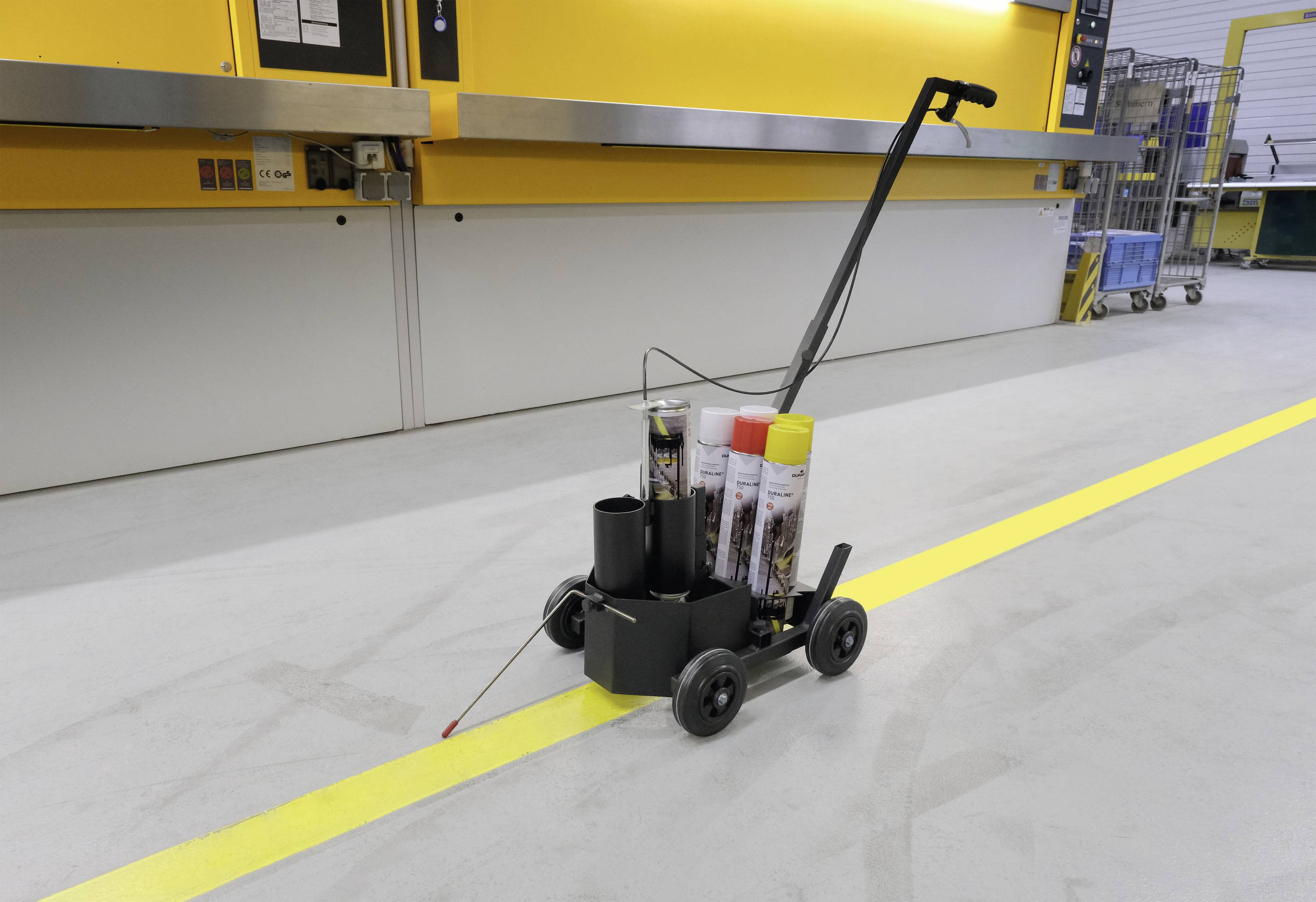 Czterokołowy wózek z uchwytem stoi na betonowej powierzchni przed żółtą ścianą. Przewozi różne pojemniki, w tym dwa ze sprayem do malowania.