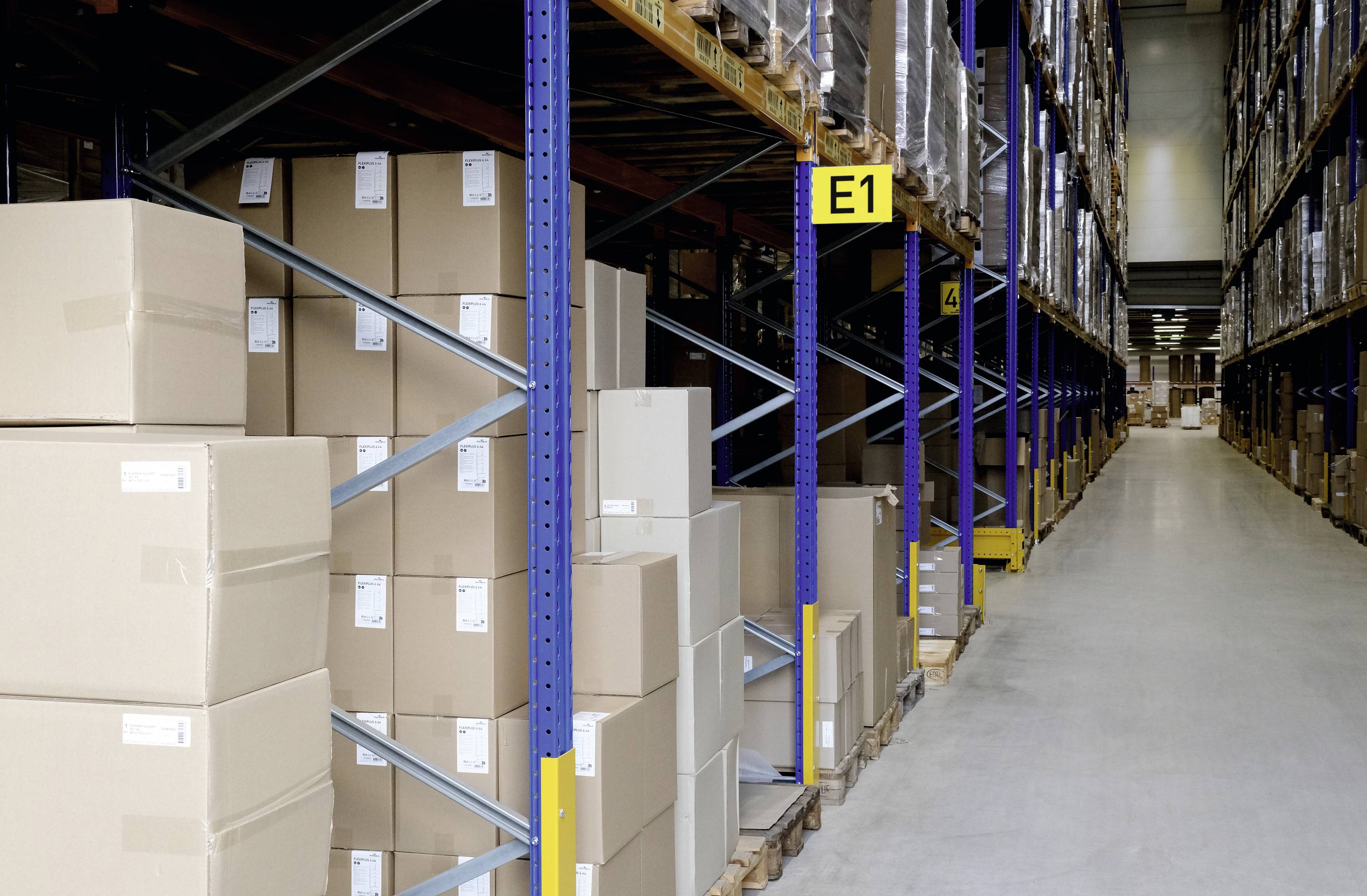 Rzędy kartonów w magazynie wysokiego składowania. Wysokie regały wypełnione stosami pudełek, które obramowują wąski korytarz.