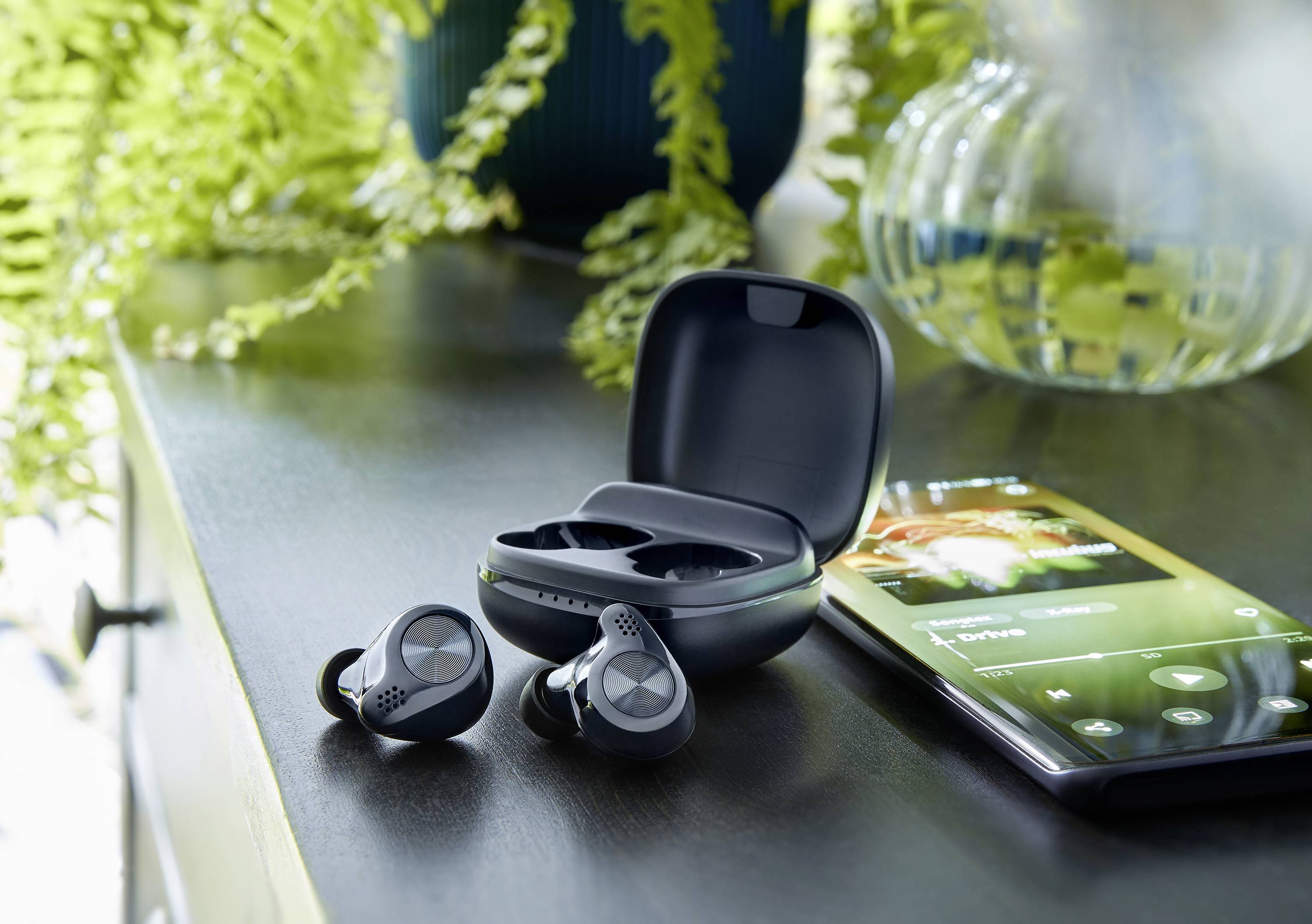 Para bezprzewodowych słuchawek dousznych leży obok otwartego etui ładującego na stole. Obok znajduje się smartfon z aplikacją muzyczną.