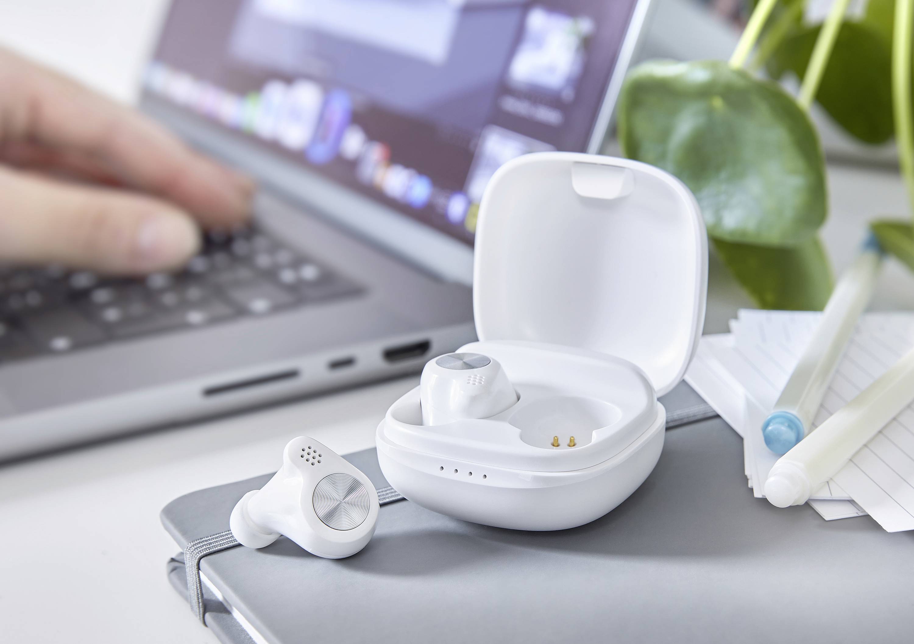 Bezprzewodowe słuchawki douszne leżą w otwartym etui ładującym na laptopie. W tle widać osobę piszącą na klawiaturze.