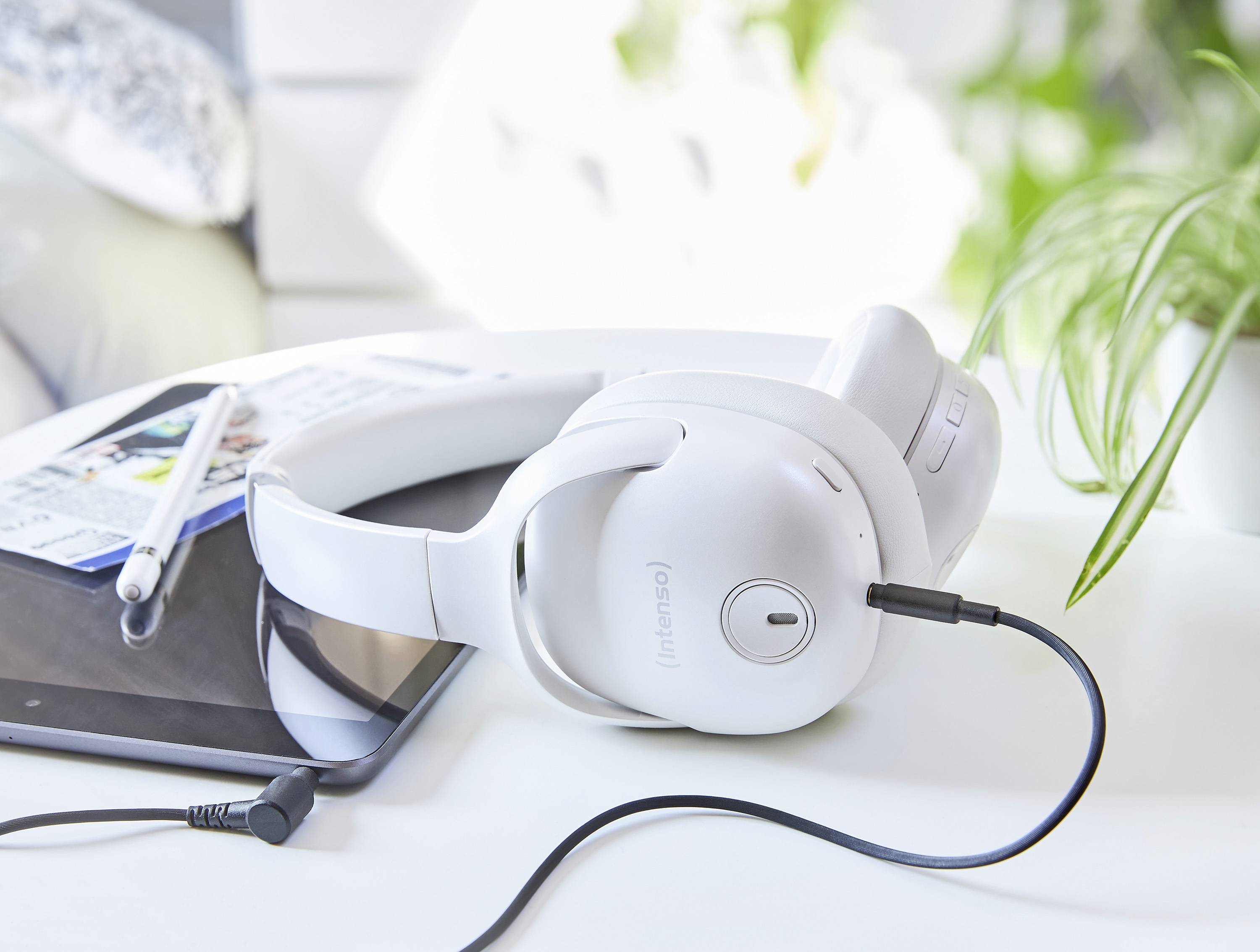 Białe słuchawki przewodowe leżą na tablecie, obok niego znajduje się długopis. Jasne, nowoczesne otoczenie z roślinami w tle.
