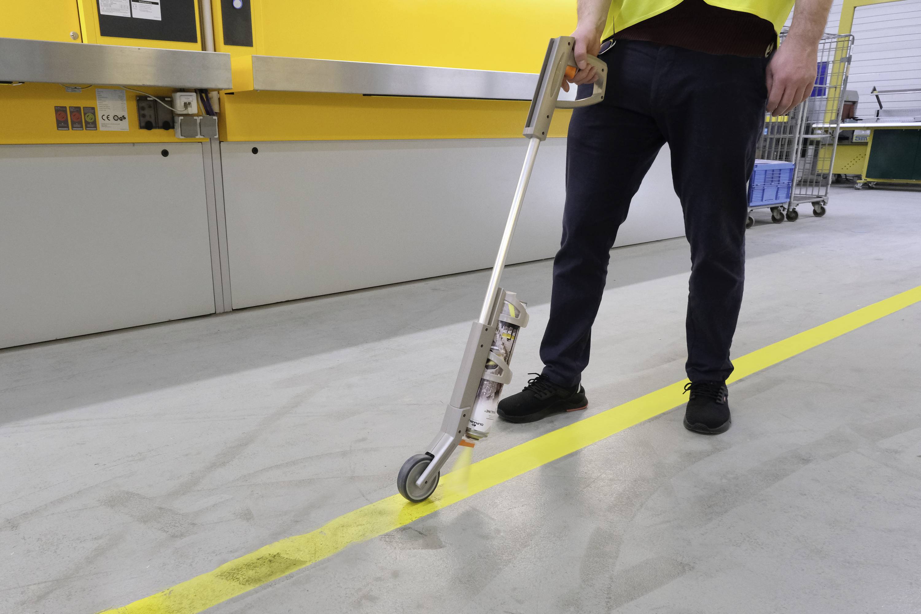 Osoba rysuje żółtą linię na szarym podłożu przy pomocy urządzenia. Na tle widoczna jest żółta ściana.