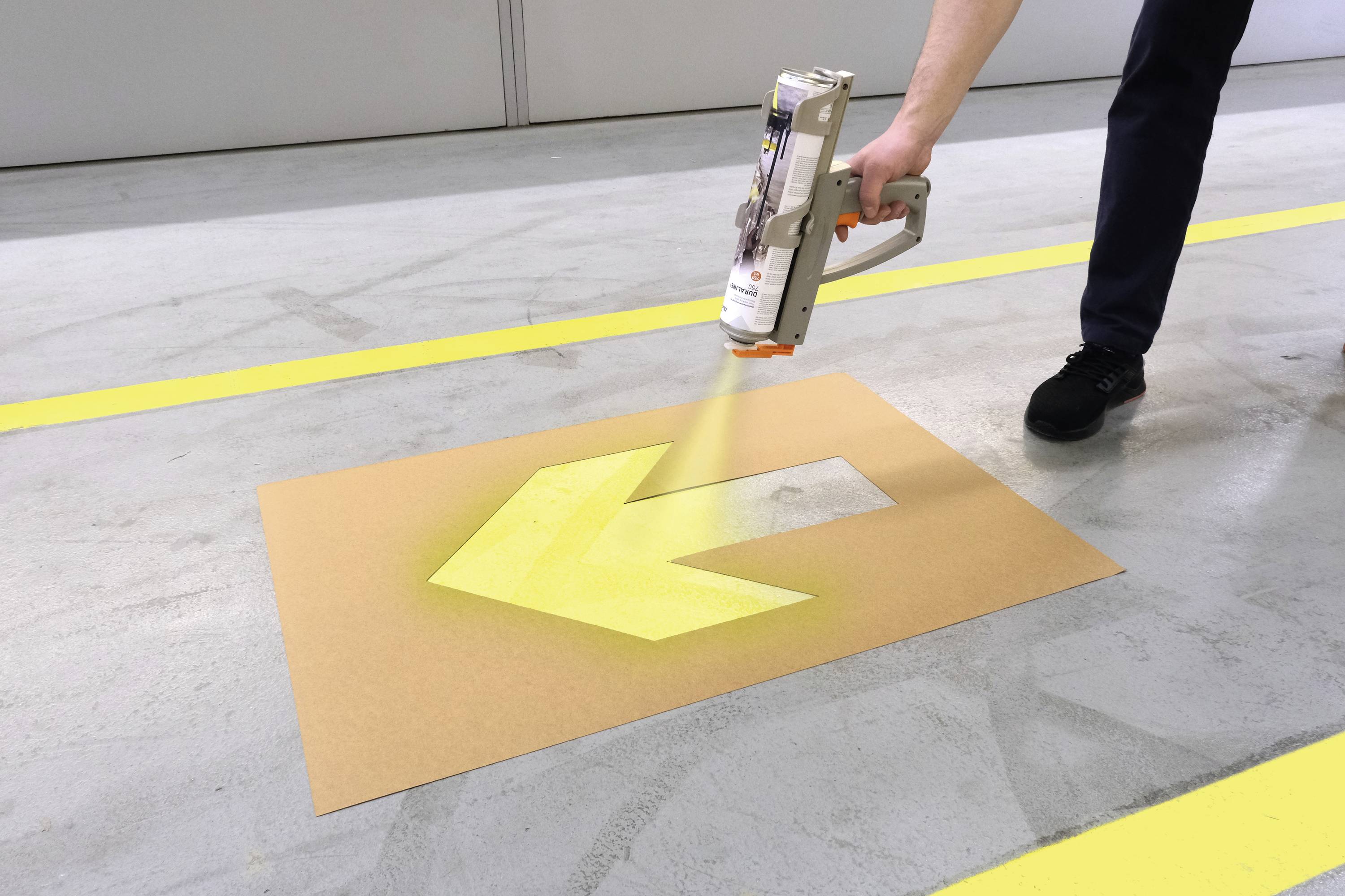 Osoba sprayuje żółtą strzałkę na podłodze hali magazynowej. Strzałka skierowana jest w lewo i służy jako wskazówka kierunkowa.