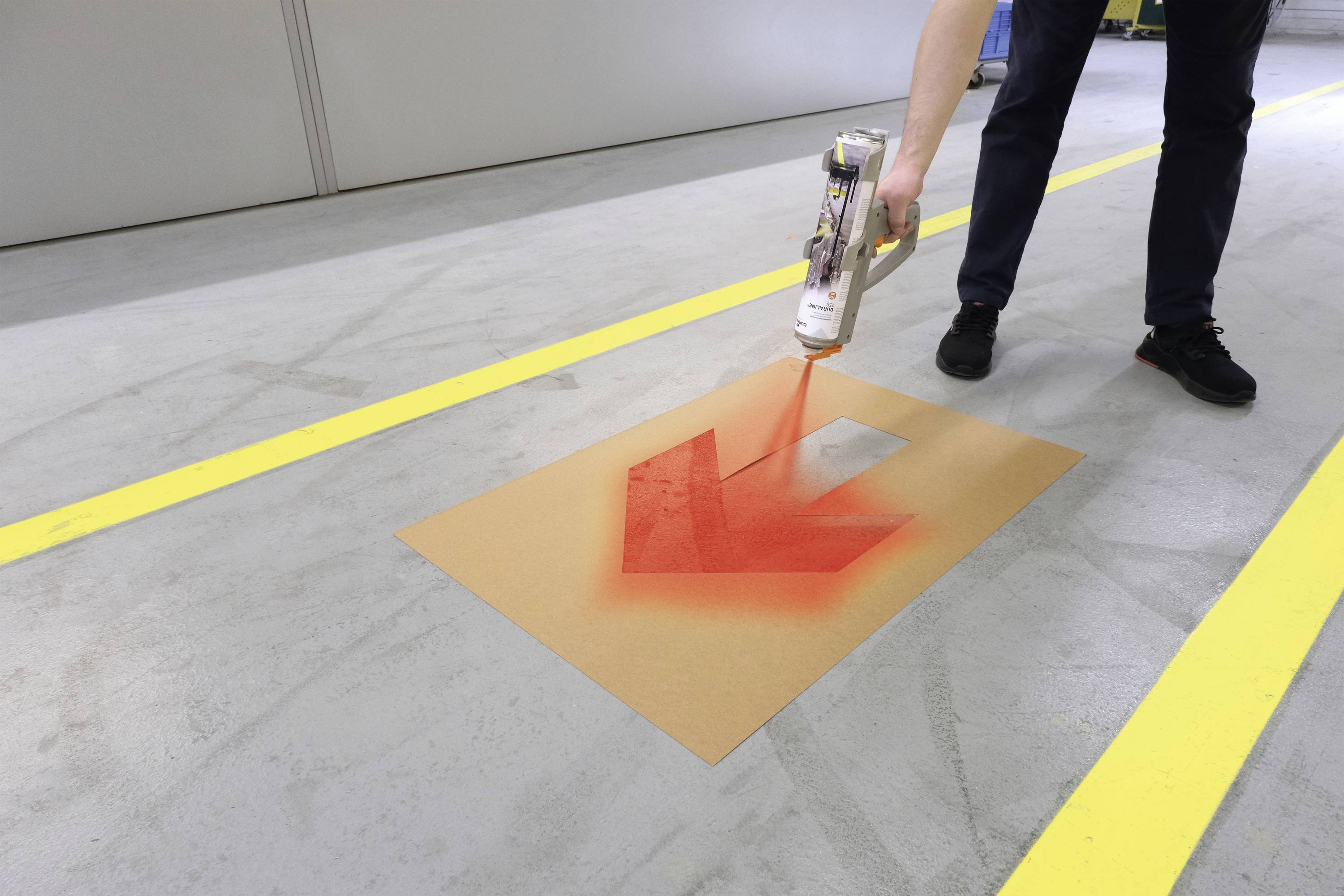 Osoba maluje duży czerwony strzałę na podłodze w warsztacie lub hali magazynowej, aby wskazać kierunek. Żółte linie oznaczają obszar.