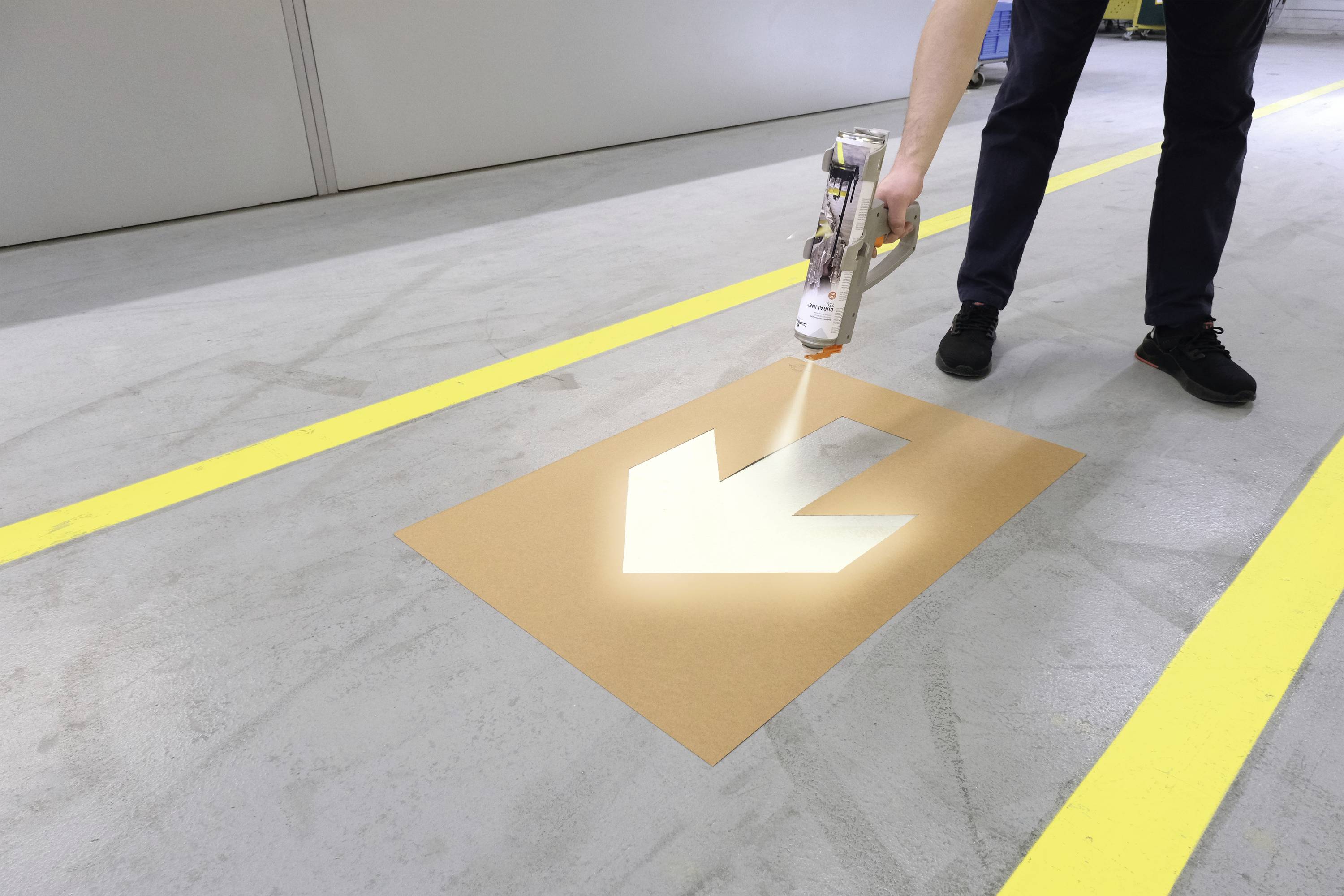 Osoba maluje biały strzałę na podłodze hali produkcyjnej za pomocą szablonu i farby w sprayu.