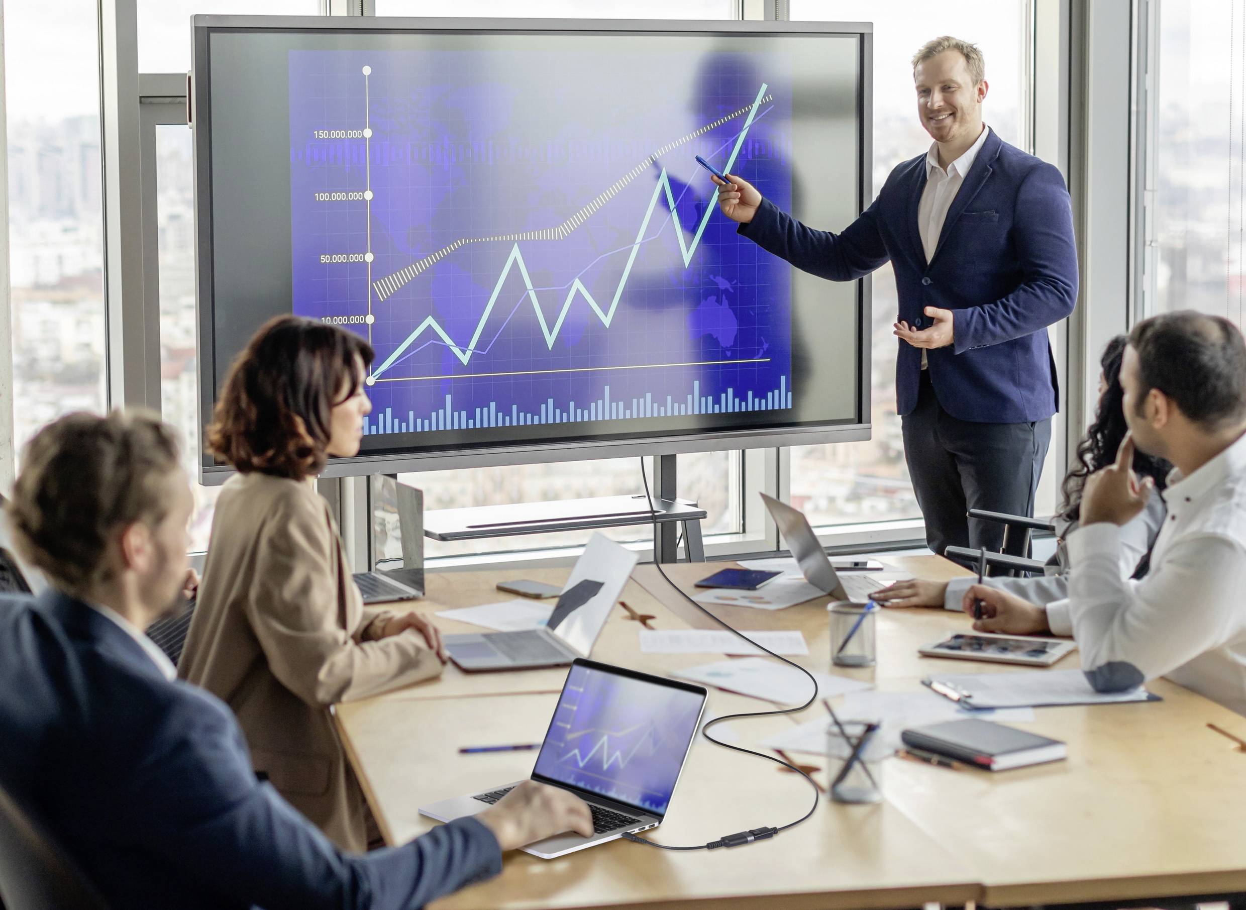 Biznesmen prezentuje wykres z rosnącymi trendami na ekranie, podczas gdy cztery osoby uważnie słuchają w sali konferencyjnej.