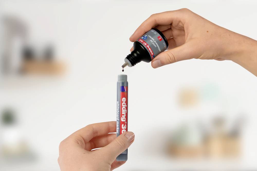 Ręka napełnia końcówkę markera edding 3000 tuszem z małej buteleczki. W tle widać nieostre obiekty.