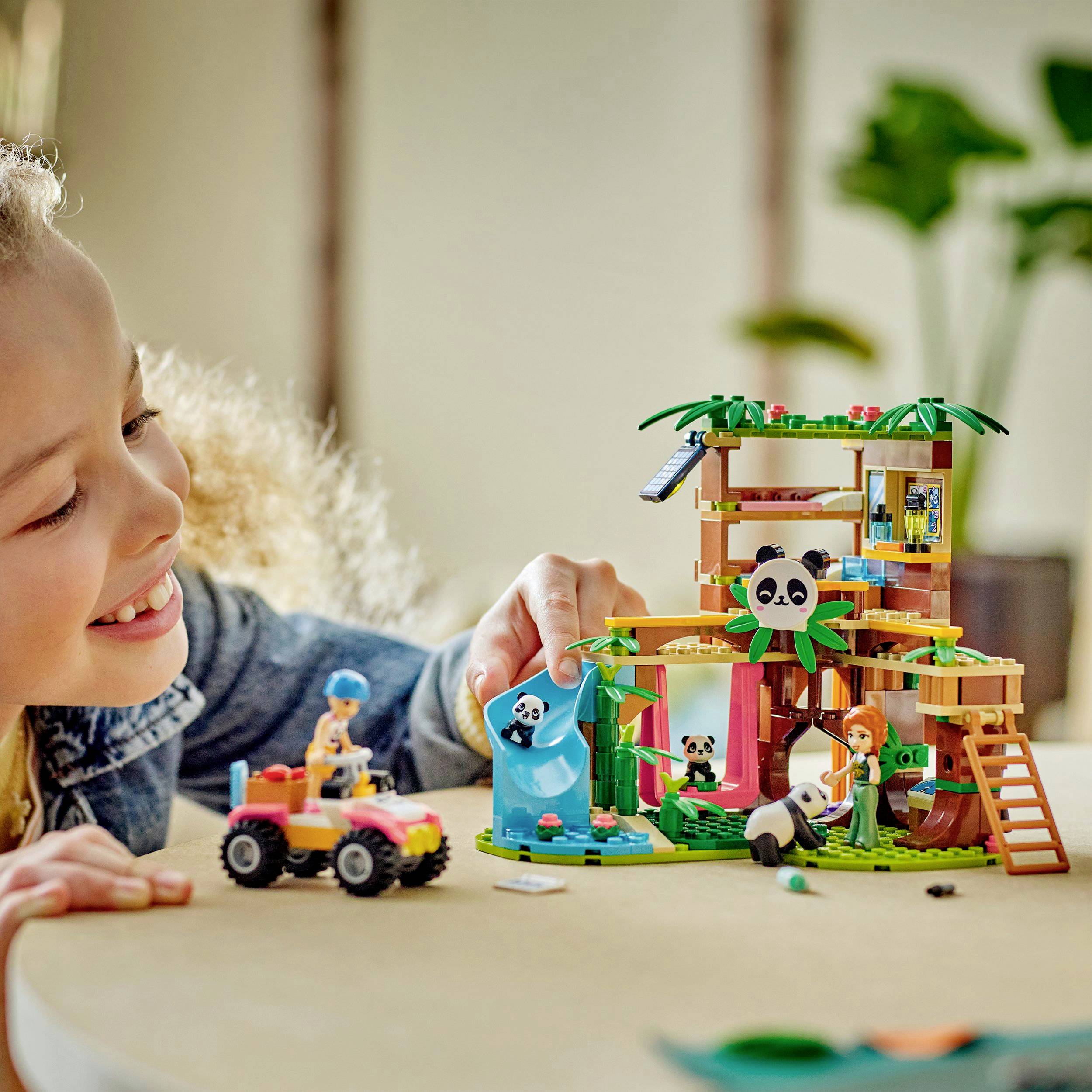 Uśmiechnięte dziecko bawi się zestawem Lego przedstawiającym scenę dżungli z pandami. W tle widać rośliny.
