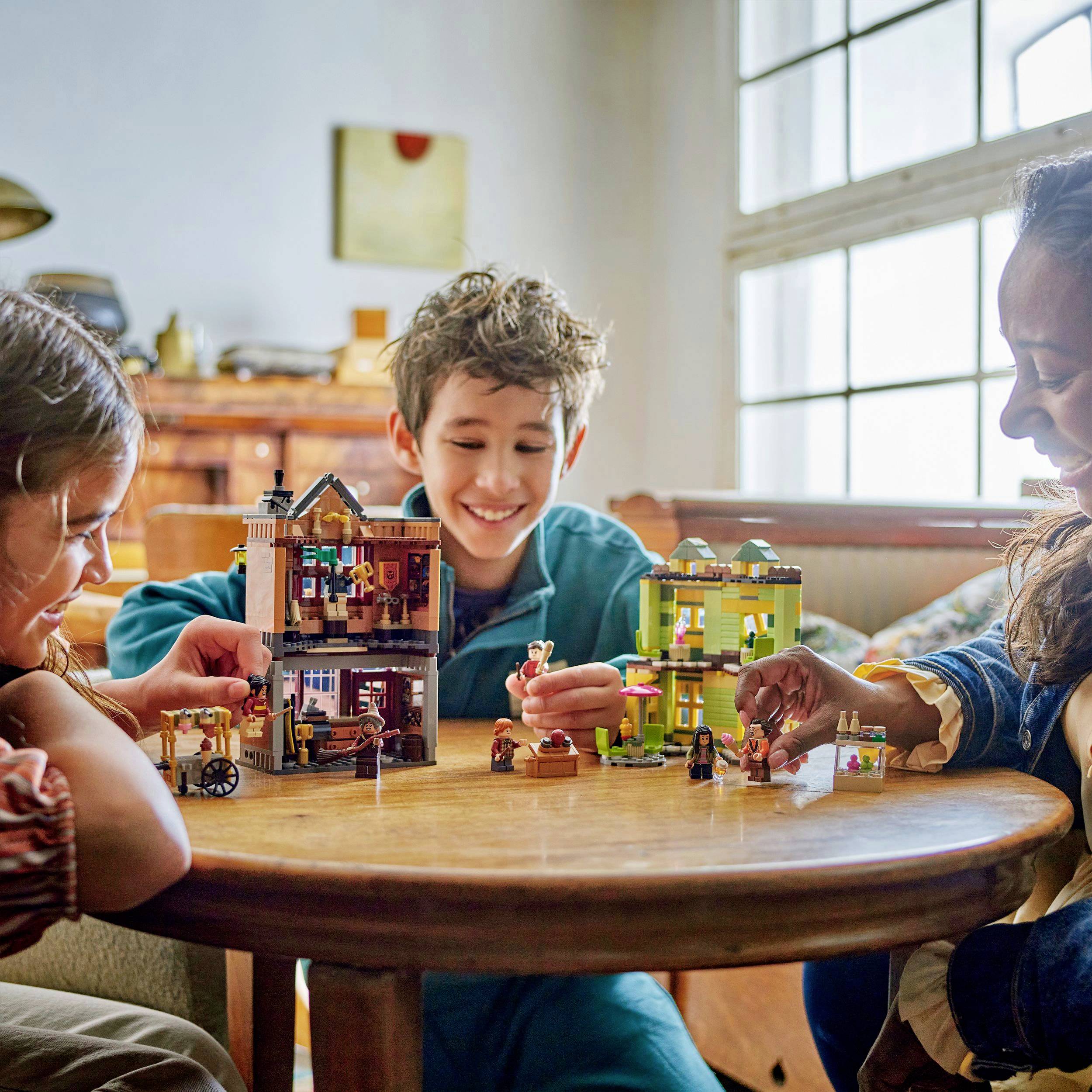 Troje dzieci siedzi przy okrągłym stole i bawi się obszernym zestawem Lego, który zawiera domki i minifigurki. Wyglądają na szczęśliwe i skoncentrowane.