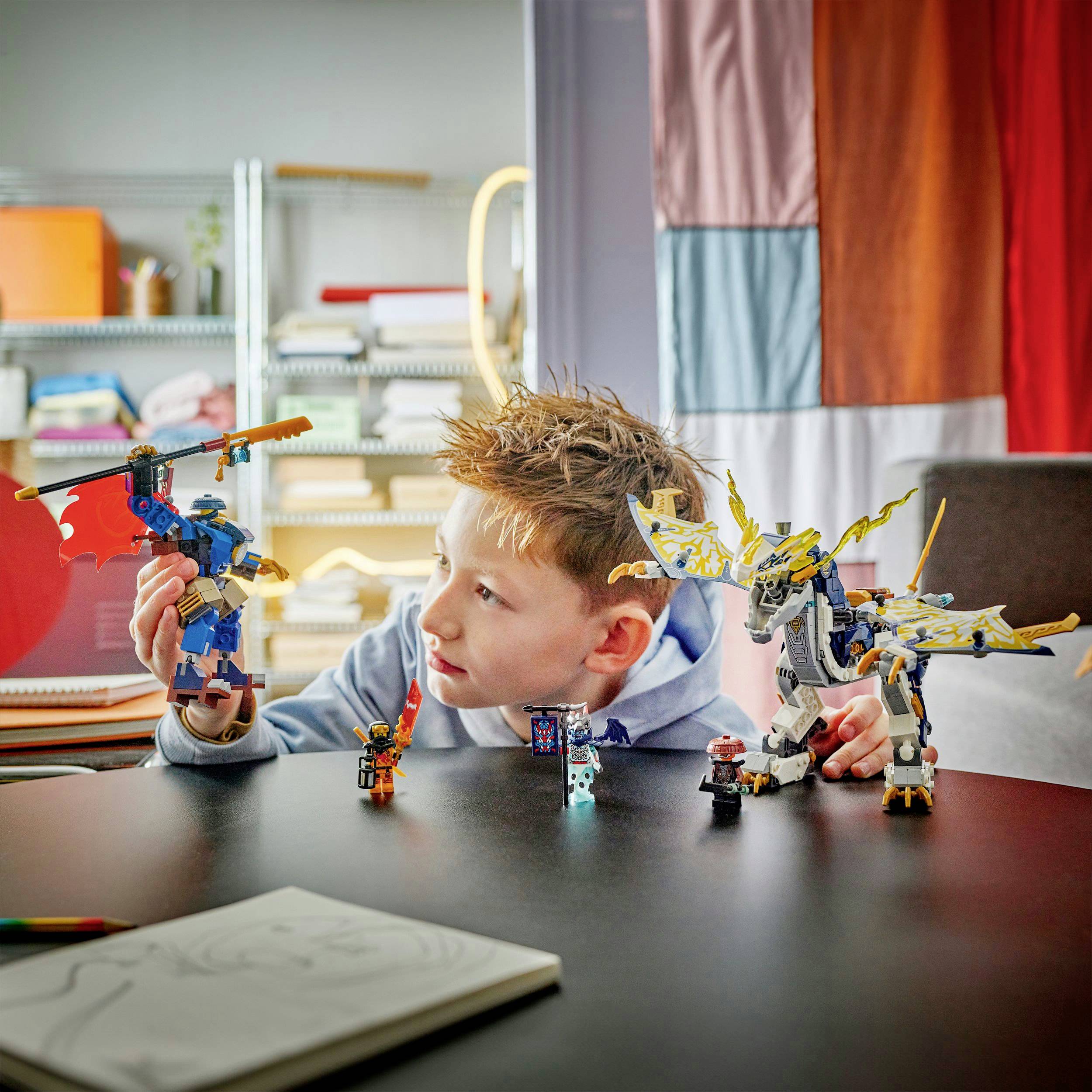 Dziecko bawi się dwoma kolorowymi zabawkowymi robotami na stole w jasnym pokoju. W tle widoczne są półki.