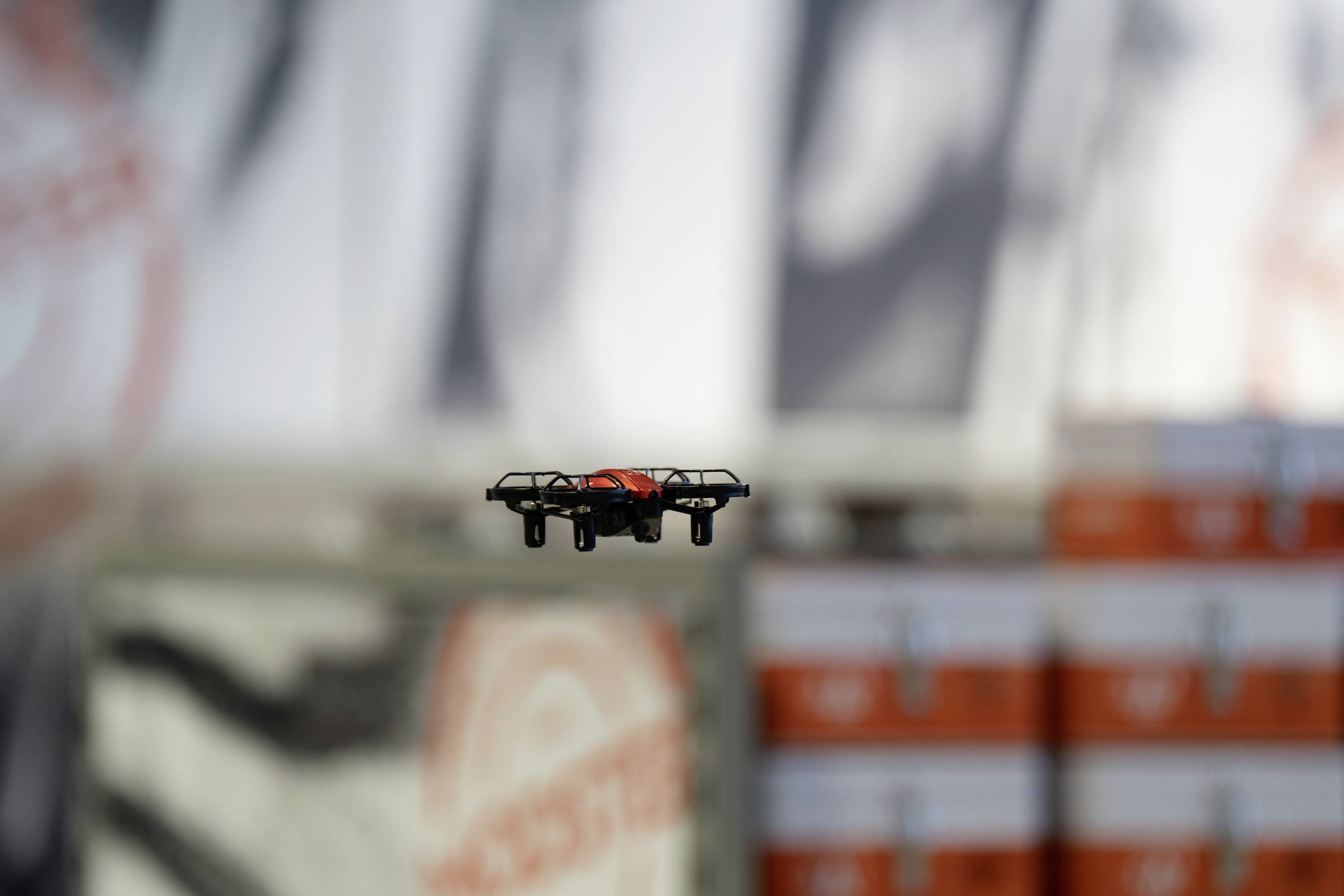 Mała dronka unosi się w pomieszczeniu na tle rozmytych pomarańczowo-białych kartonów.