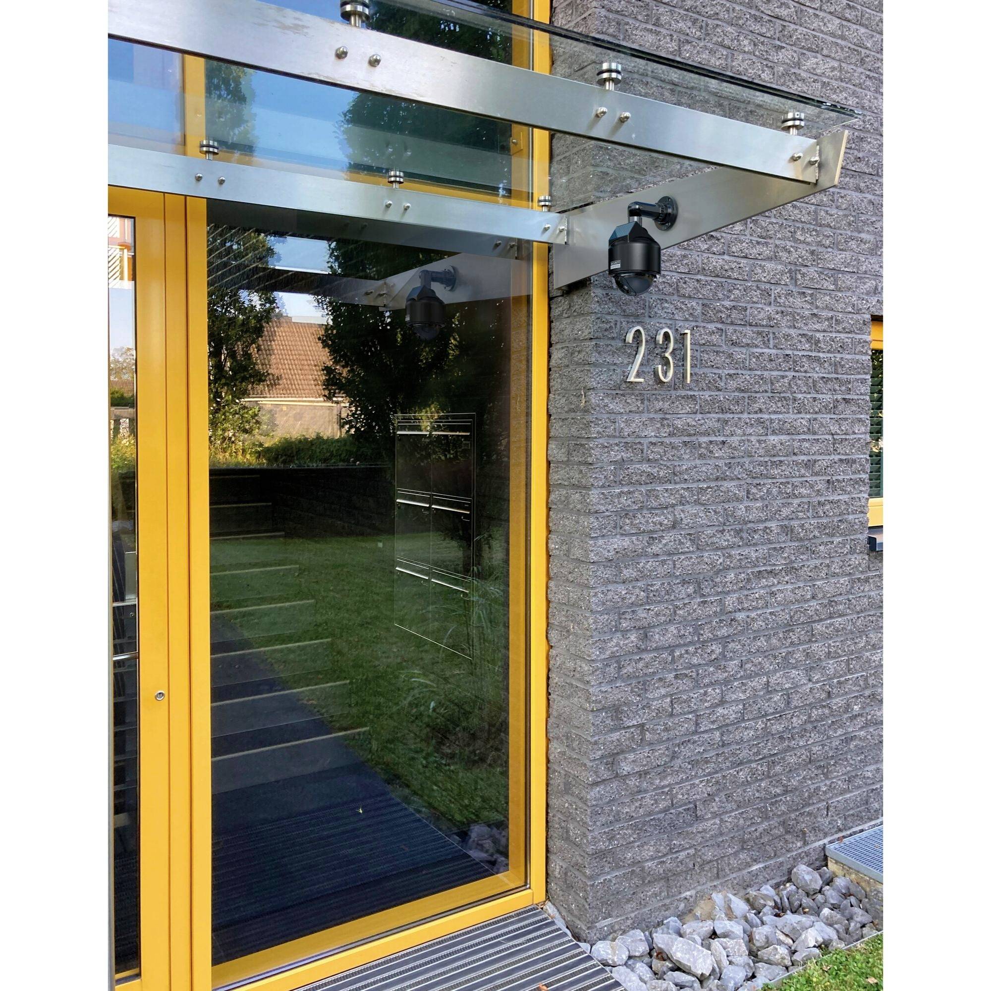 Wejście do nowoczesnego budynku ze szklanym zadaszeniem, żółtą ramą drzwi i numerem domu '231'. Widoczny spokojny, zielony dziedziniec.