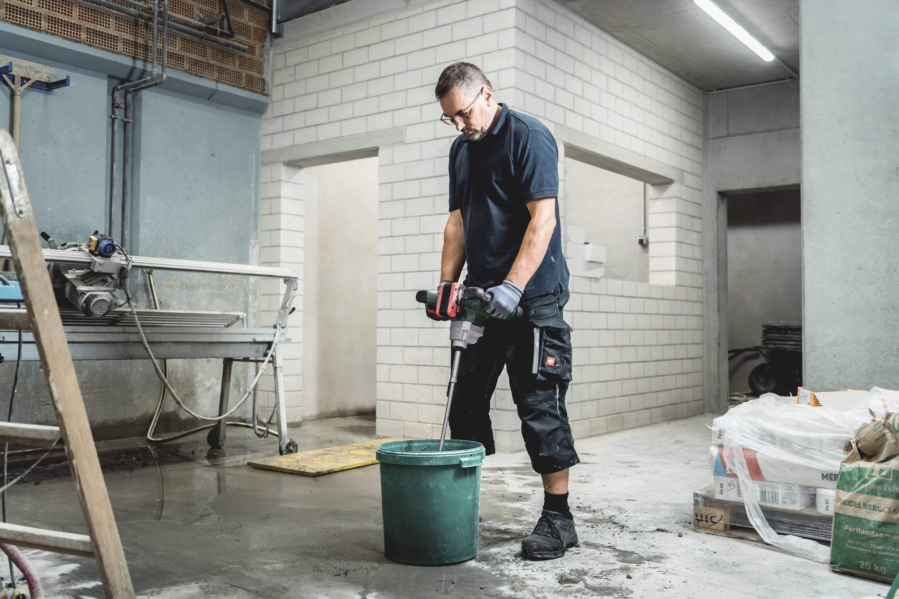 Mężczyzna w odzieży roboczej miesza materiał elektrycznym mieszadłem w wiadrze na placu budowy.