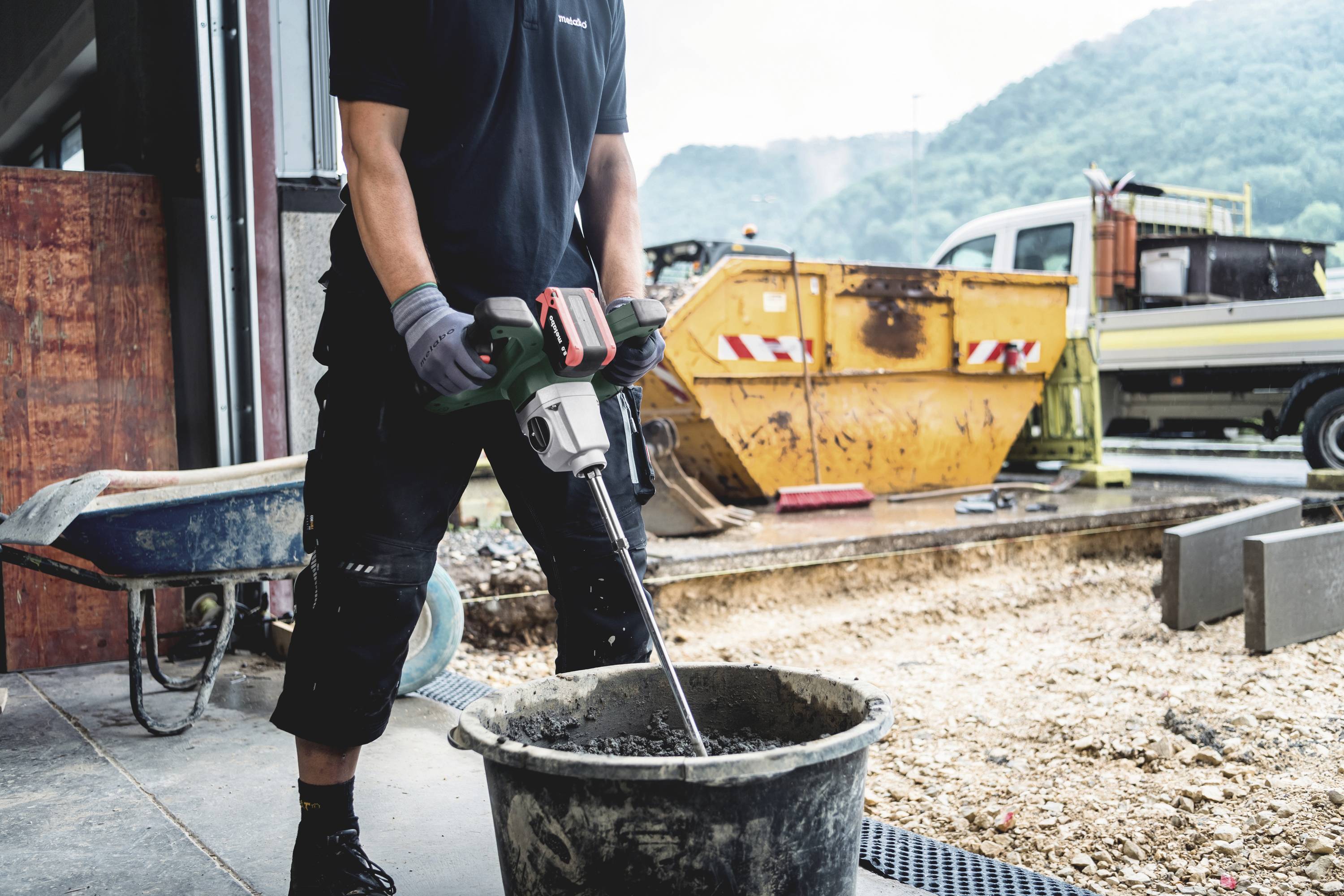 Robotnik miesza beton za pomocą mieszalnika na placu budowy, w tle widoczne są kontenery i ciężarówki.