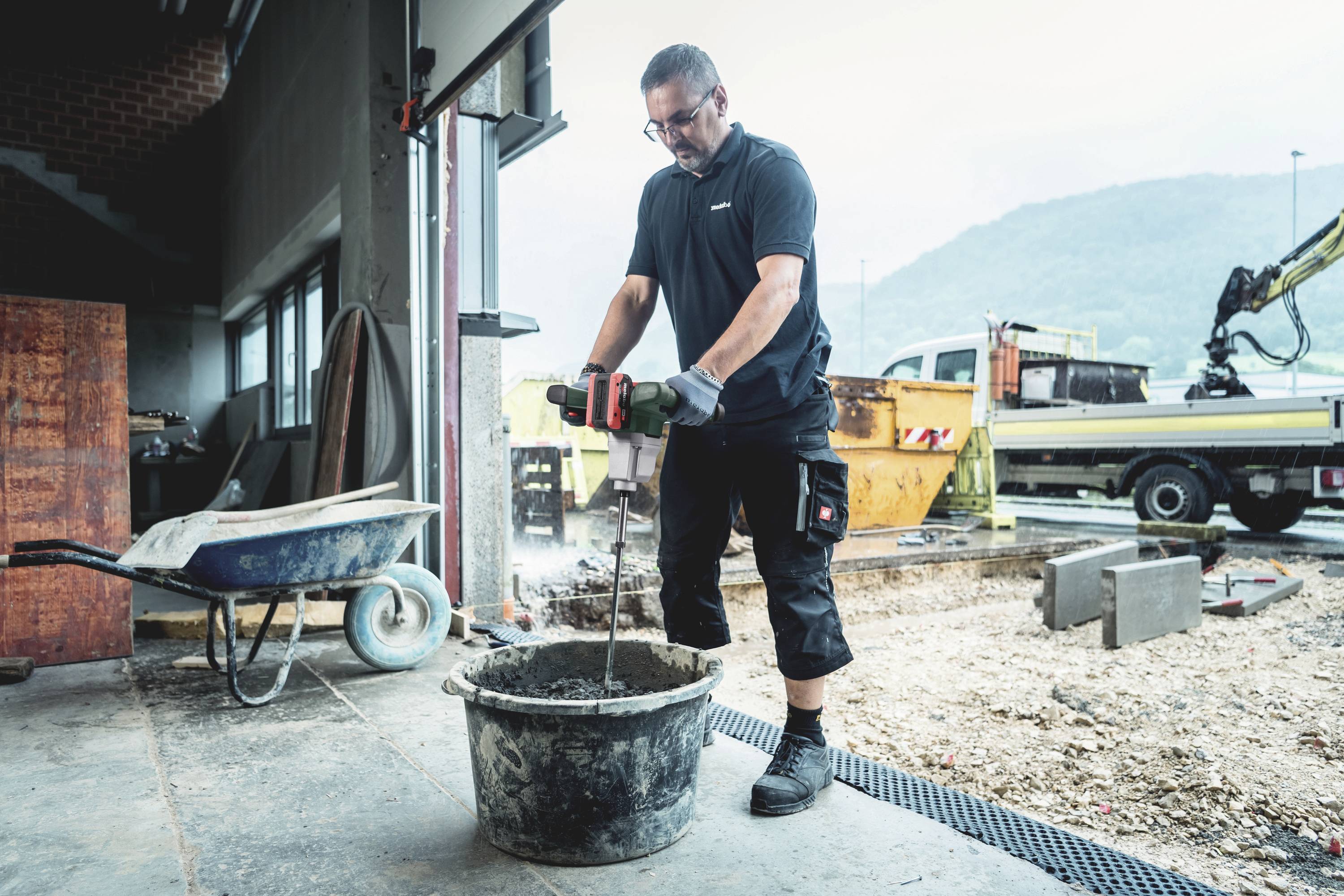 Mężczyzna miesza cement elektrycznym narzędziem w wiadrze budowlanym w warsztacie. W tle widoczne są pojazdy budowlane.