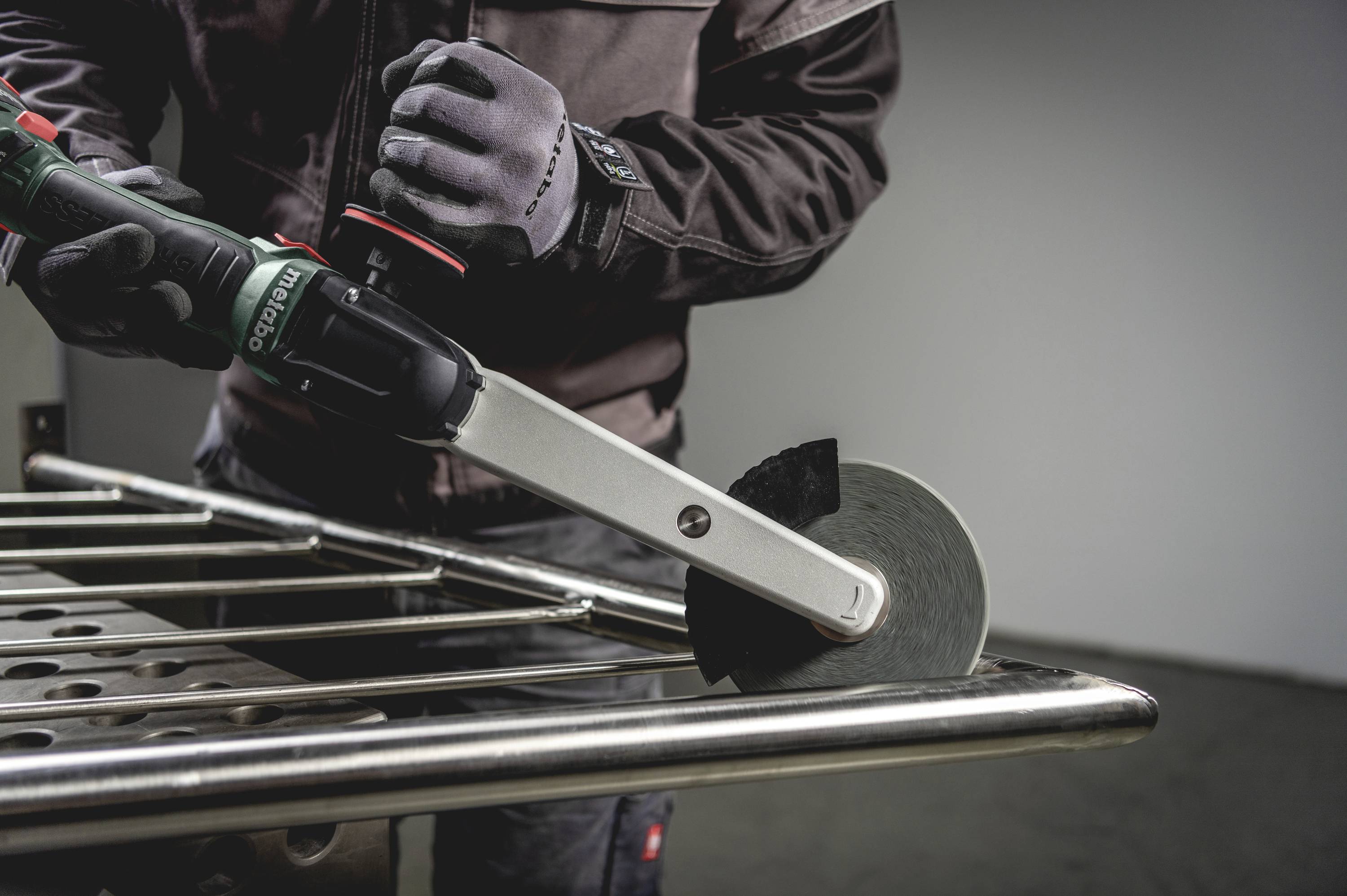 Osoba pracuje z polerką kątową przy metalowej poręczy, ubrana jest w odzież ochronną i rękawice.