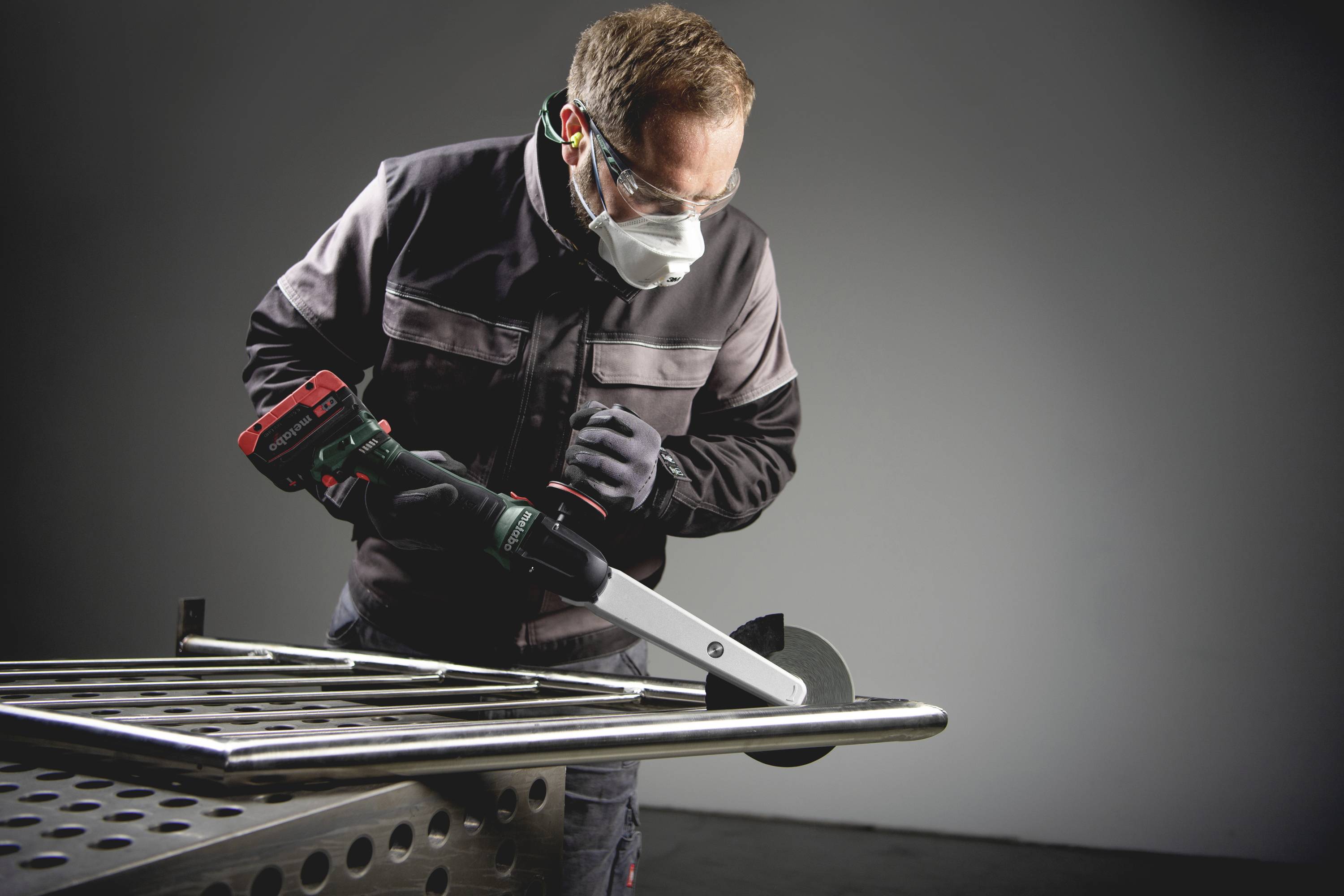 Mężczyzna w okularach ochronnych i masce tnie metalową rurę elektronarzędziem w warunkach warsztatowych.