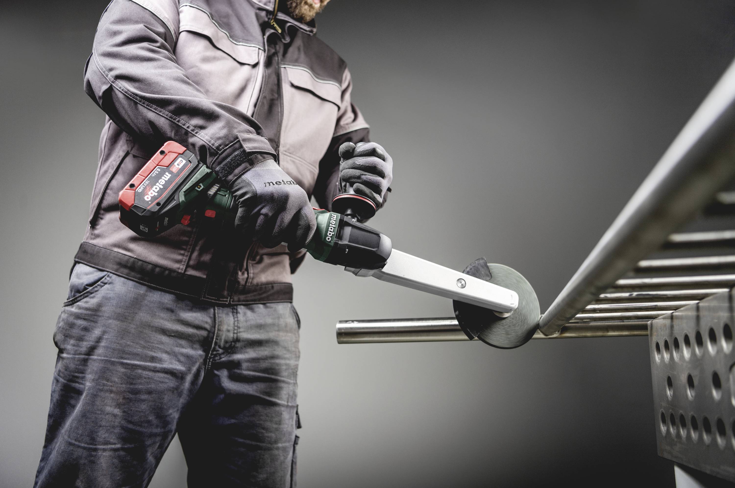 Robotnik szlifuje rurę metalową przy pomocy elektrycznej szlifierki kątowej. Jest ubrany w odzież ochronną i rękawice.