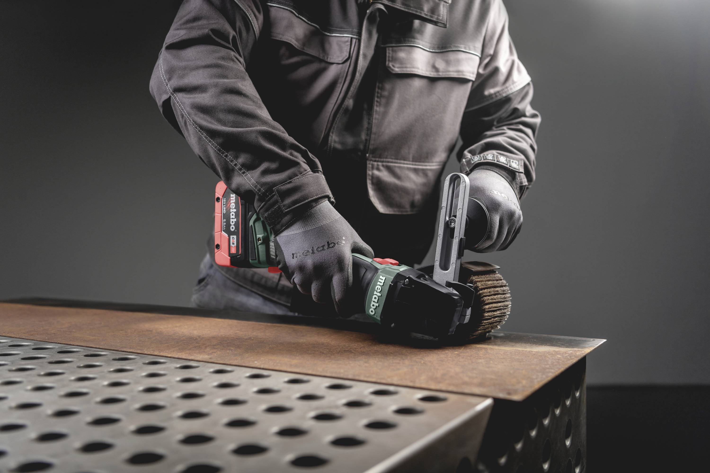 Osoba szlifuje metalową płytę elektronarzędziem na stole z wzorem otworów, nosząc przy tym rękawice ochronne.