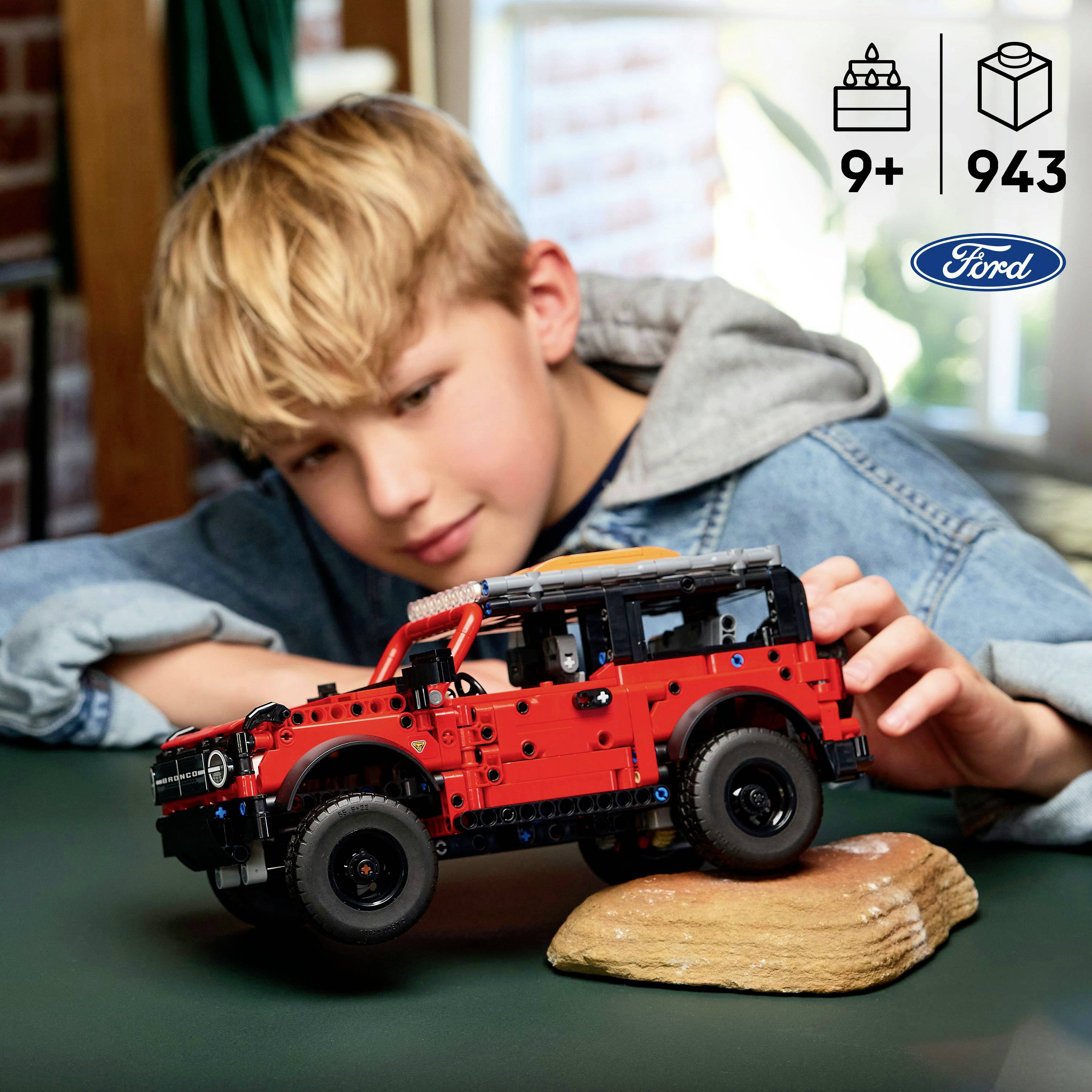 Dziecko bawi się czerwonym modelem LEGO Forda Bronco na stole. Ikony na pudełku sugerują, że zestaw jest odpowiedni dla dzieci w wieku 9 lat i starszych.
