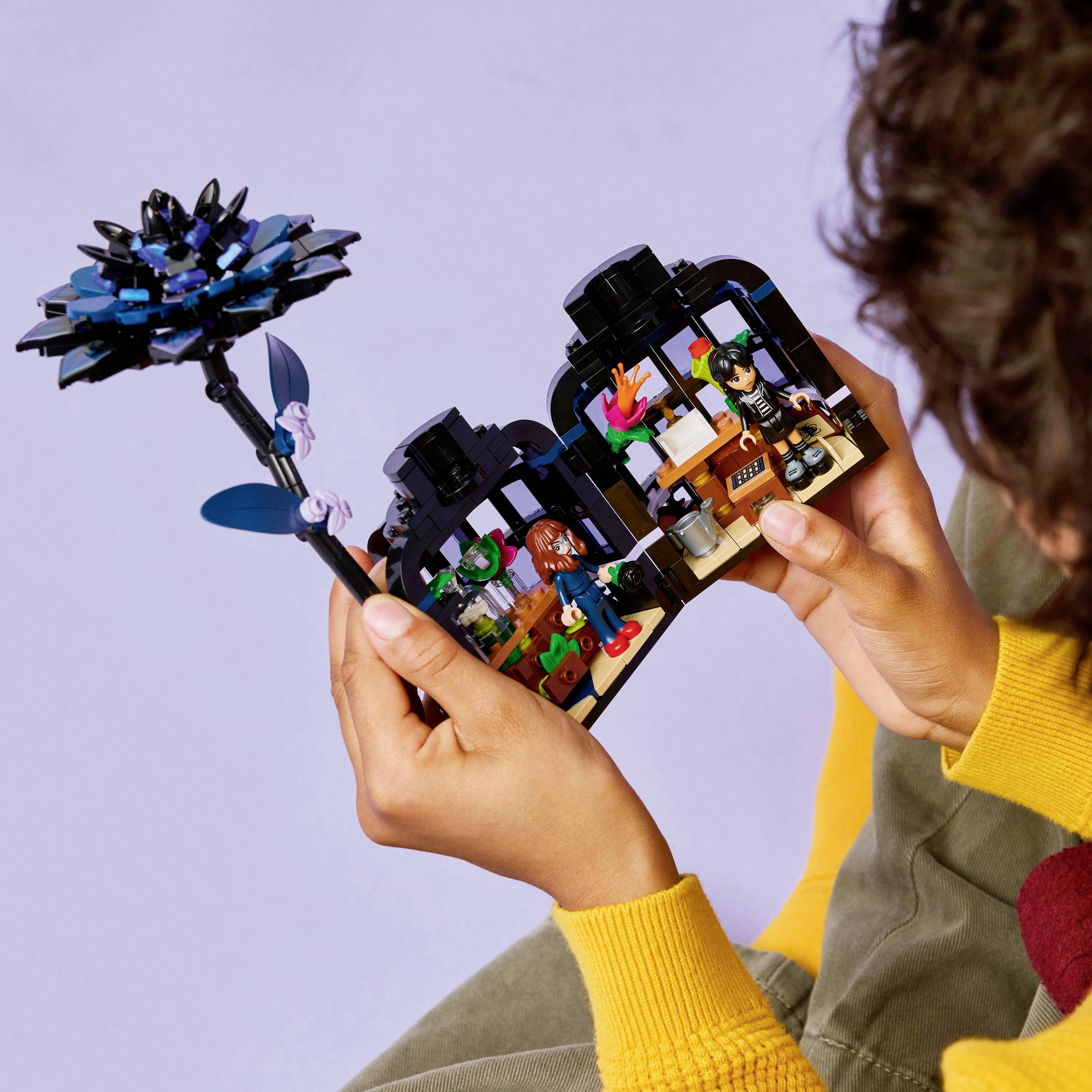 Dwie ręce trzymające model kwiatka z klocków Lego oraz otwartą miniaturową scenę pokoju z małymi figurkami, ilustrujące kreatywne budowanie z klocków Lego.