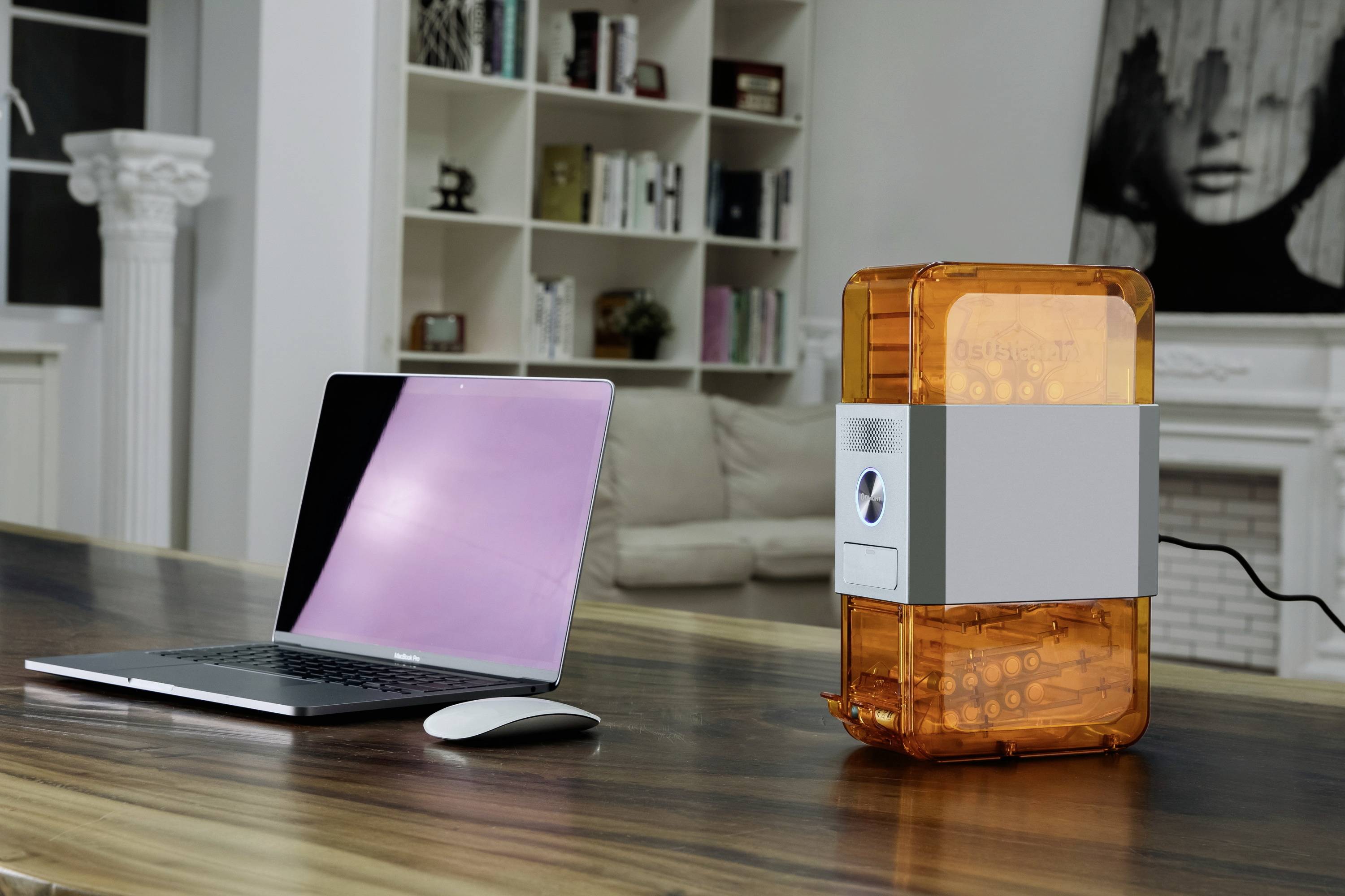 Laptop i pomarańczowy oczyszczacz powietrza stoją na stole w nowoczesnym salonie, a w tle widać regał z książkami.