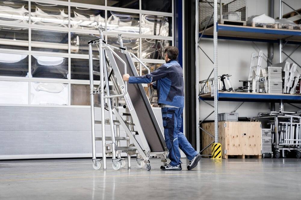 Osoba przesuwa przenośną aluminiową drabinę w magazynie. W tle widoczne są regały z częściami metalowymi.