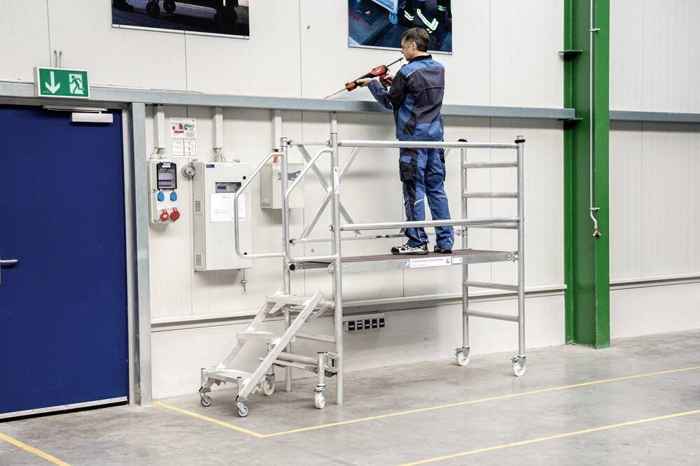 Osoba w stroju roboczym stoi na rusztowaniu jezdnym w hali przemysłowej i pracuje narzędziem przy ścianie.