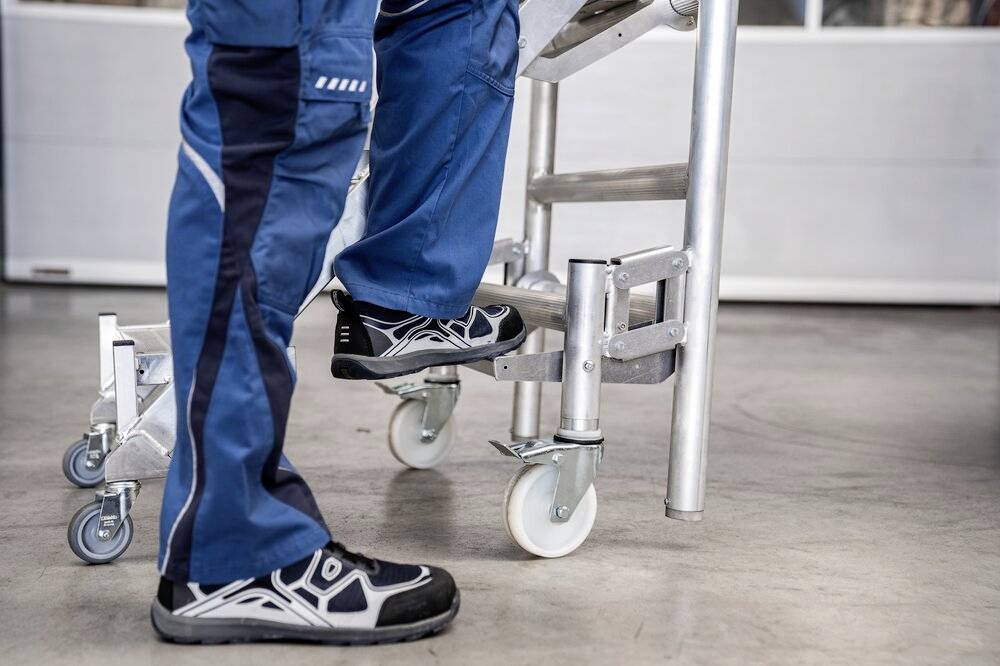 Osoba w niebieskich spodniach roboczych wchodzi na drabinę na kółkach. Podłoga jest szara i gładka, co wskazuje na środowisko warsztatu.
