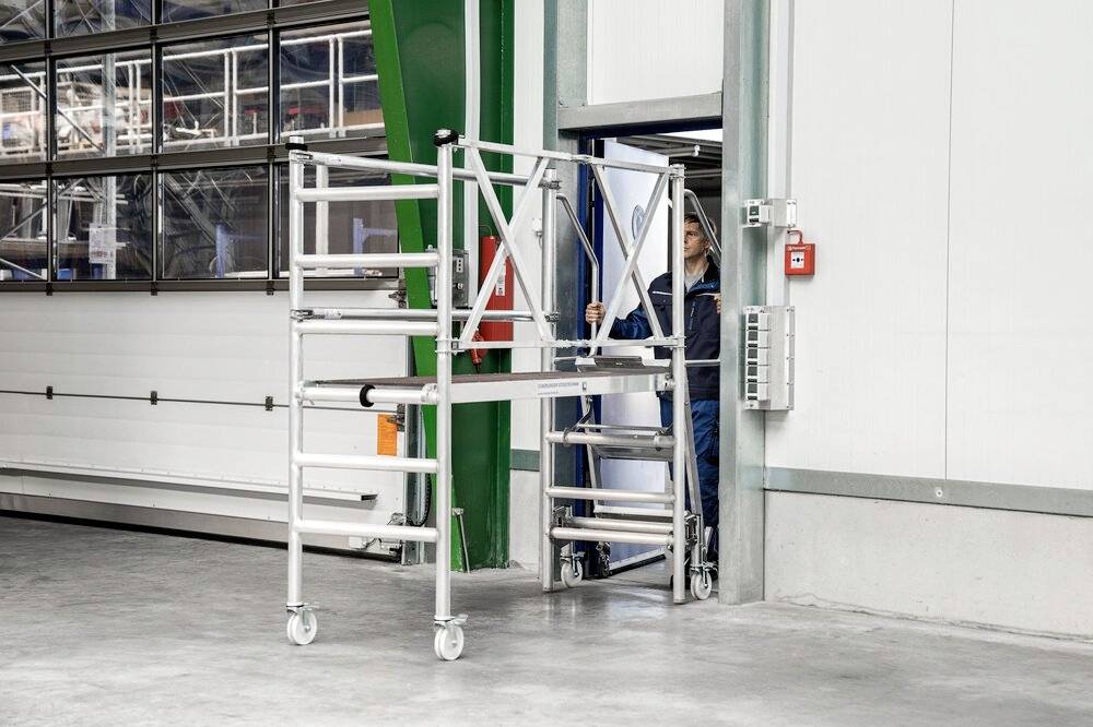 Osoba przesuwa składaną platformę aluminiową na kółkach przez drzwi w hali przemysłowej. Platforma jest częściowo złożona.