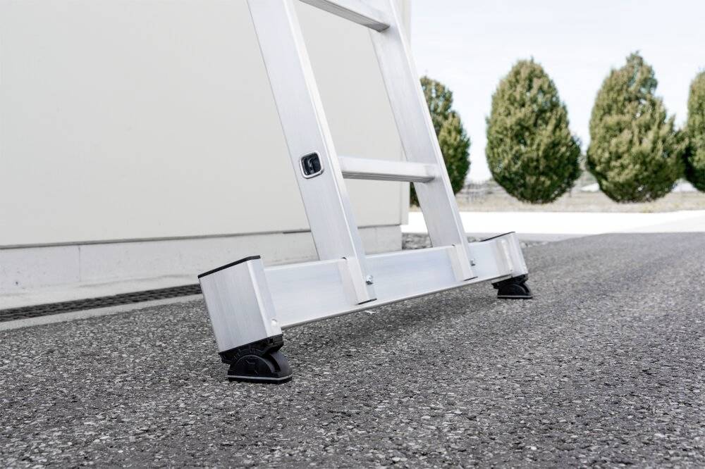Aluminiowa drabina stoi stabilnie na asfaltowej powierzchni, oparta o jasną ścianę budynku, w tle widoczne są drzewa.