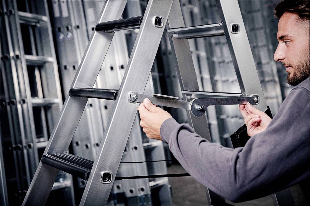 Osoba reguluje metalową drabinę w magazynie. W tle widoczne są ułożone drabiny.