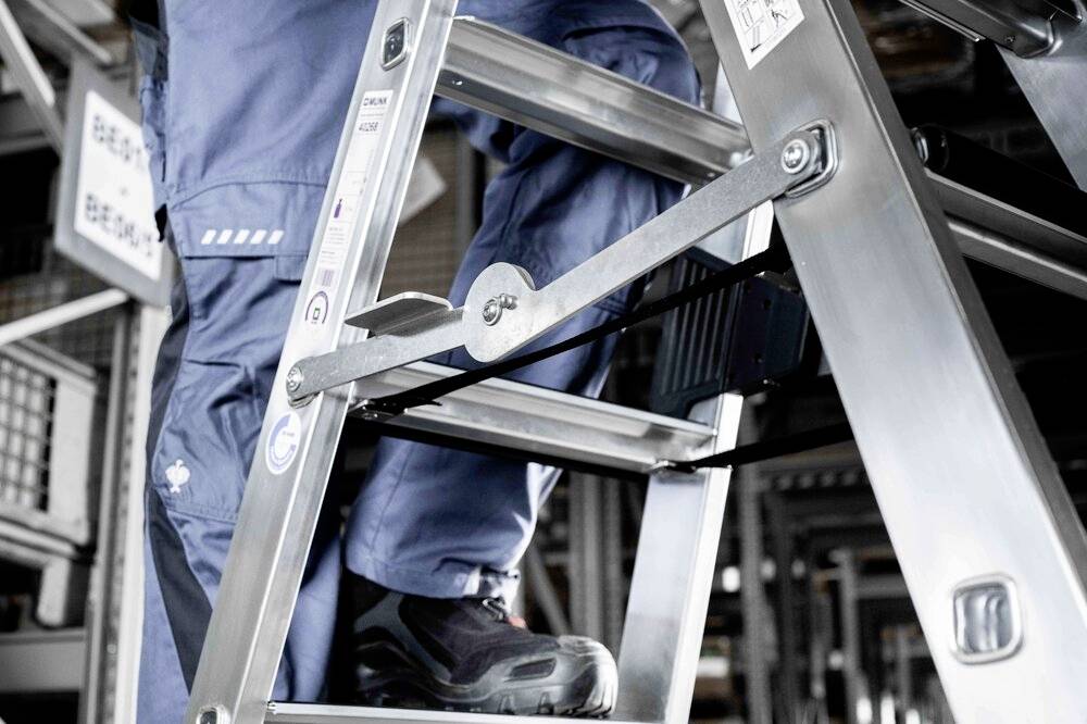 Osoba w stroju roboczym wchodzi na aluminiową drabinę w magazynie. Zdjęcie przedstawia bezpieczeństwo podczas pracy z drabiną.