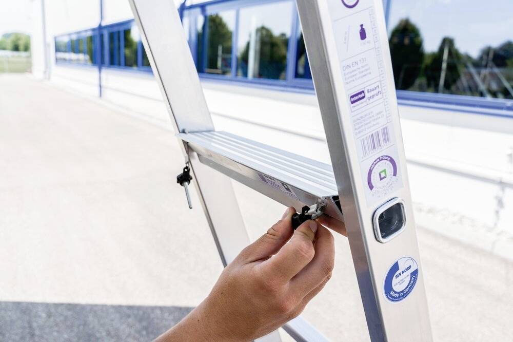 Ręka reguluje śrubę na aluminiowej drabinie na wolnym powietrzu. Drabina stoi przed nowoczesnym budynkiem ze szkła.
