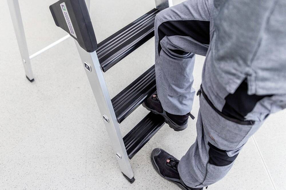 Osoba wchodzi po srebrnej drabinie składanej. Ma na sobie szare ubranie robocze i czarne buty.