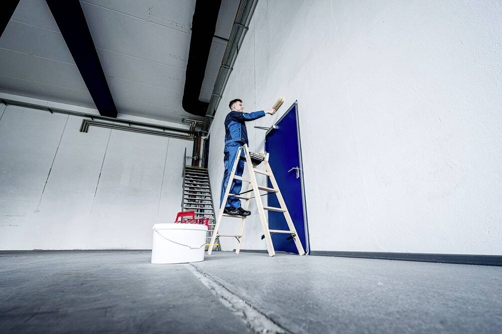 Mężczyzna stoi na drabinie i maluje niebieskie drzwi w dużym, pustym pokoju. Na podłodze stoi wiadro z farbą.