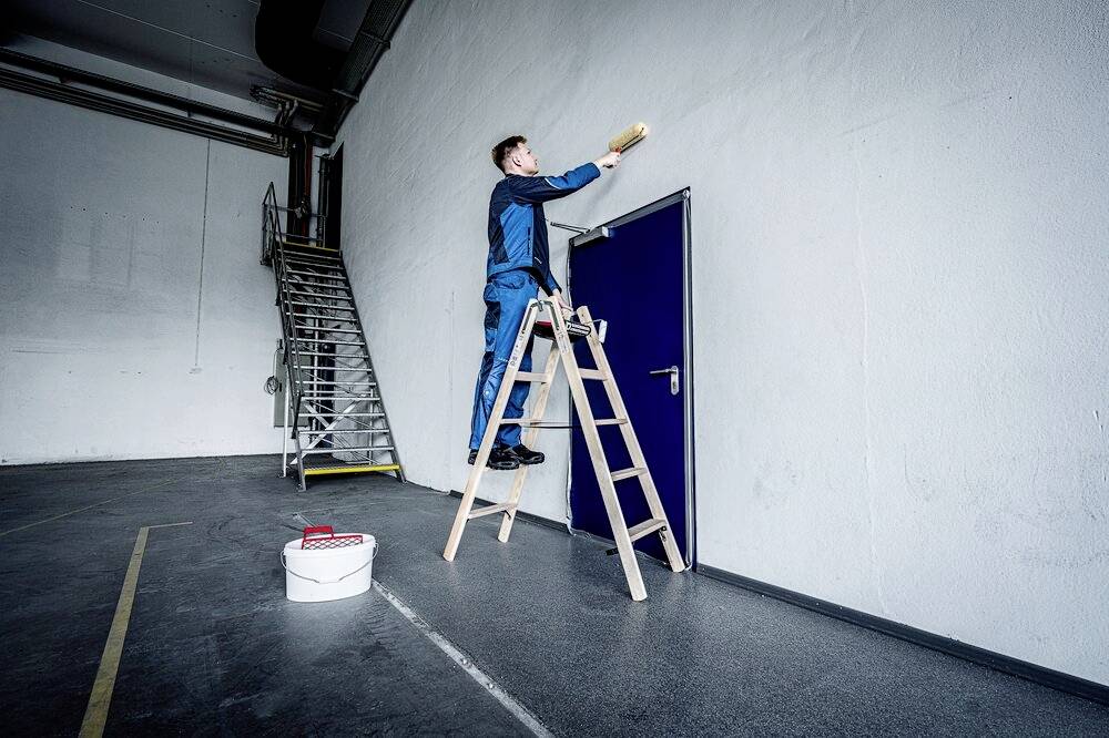 Osoba stojąca na drabinie maluje białą ścianę nad niebieskimi drzwiami. Puszka z farbą stoi na podłodze obok drabiny.