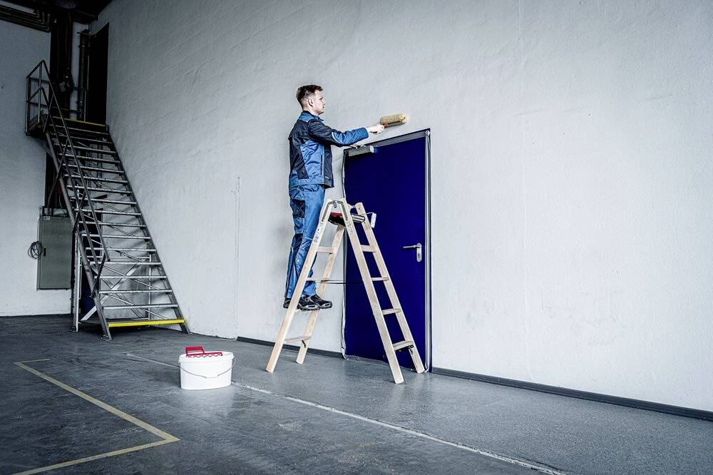 Mężczyzna w ubraniu roboczym stoi na drabinie i maluje górną połowę białej ściany nad niebieskimi drzwiami. Wiadro z farbą stoi na podłodze.