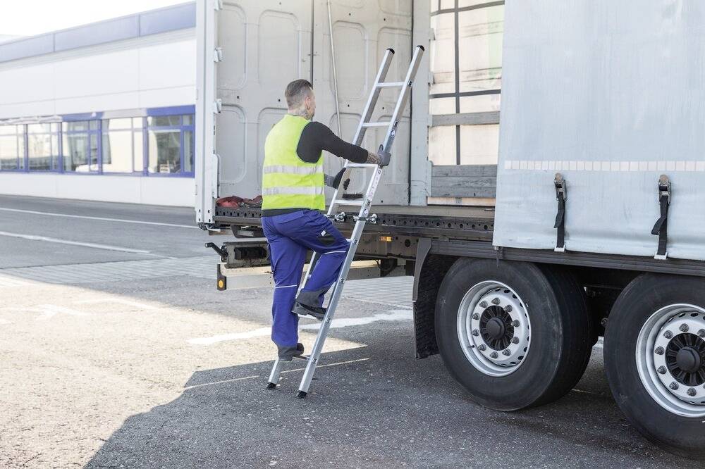 Mężczyzna w stroju roboczym wchodzi na drabinę przymocowaną do otwartego samochodu dostawczego, aby dostać się do przestrzeni ładunkowej.