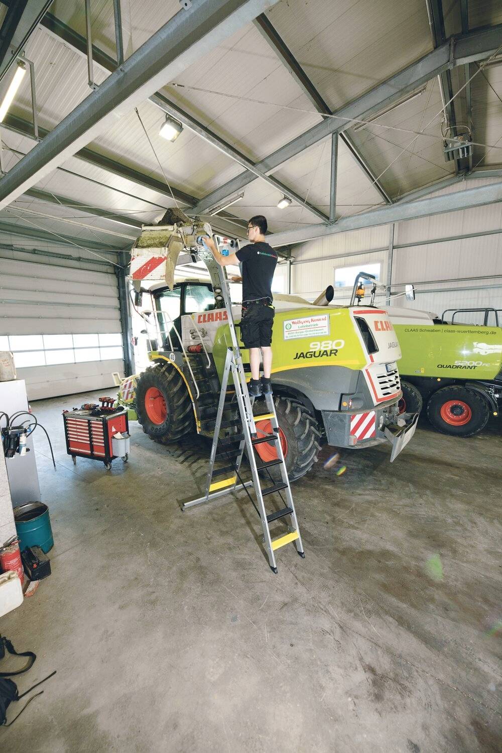 Mechanik stoi na drabinie i pracuje przy dużym traktorze w warsztacie. W tle widoczne są narzędzia.