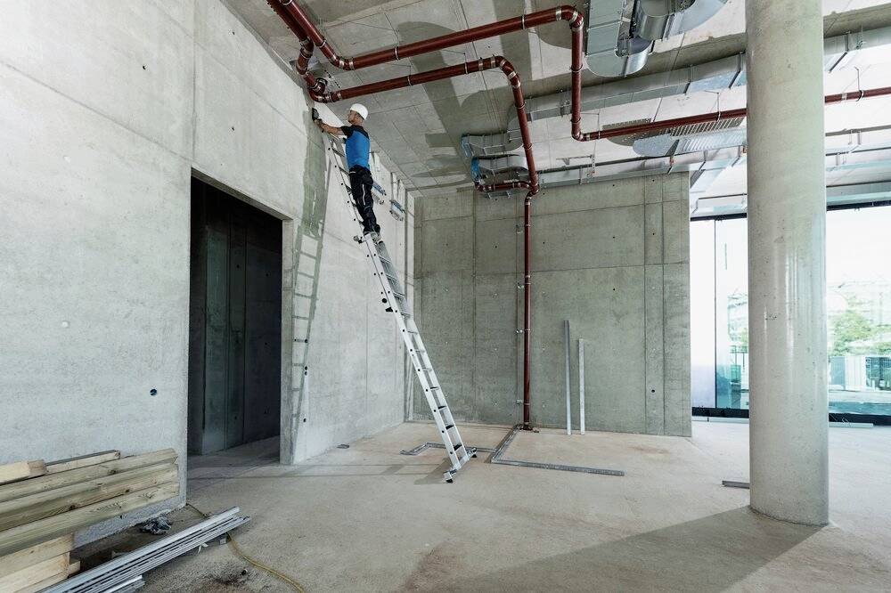 Osoba w stroju roboczym i kasku ochronnym stoi na drabinie w niedokończonym pomieszczeniu z widocznymi rurami i betonowymi ścianami.