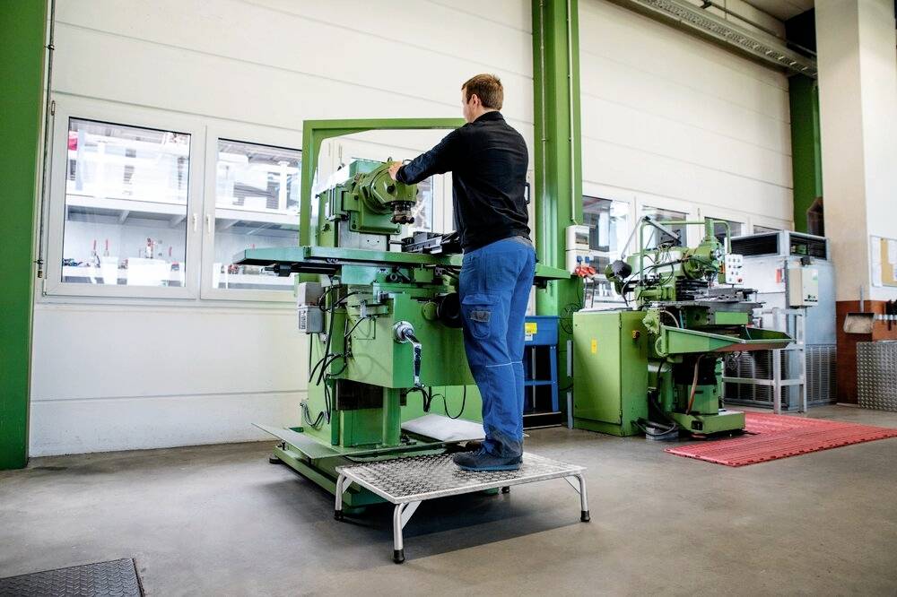 Mężczyzna w ubraniu roboczym obsługuje zieloną maszynę przemysłową w hali produkcyjnej. W tle widać kolejne maszyny.