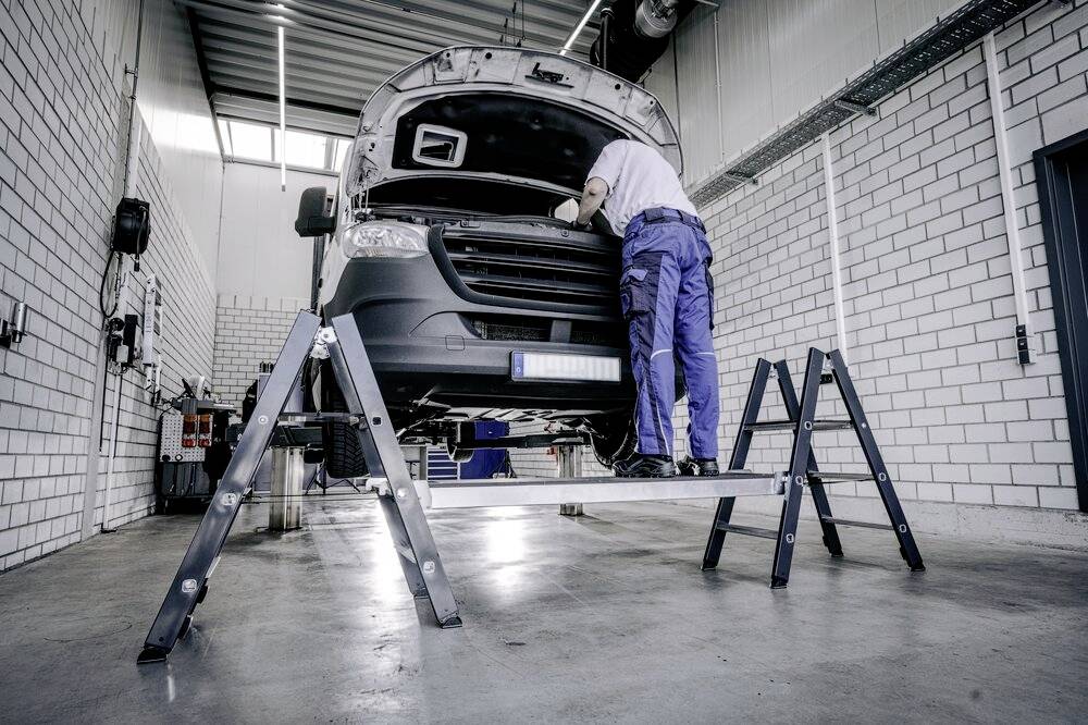 Mechanik pracuje w warsztacie na podniesionym pojeździe. Sprawdza komorę silnika. Otoczenie prezentuje przemysłowe wyposażenie.