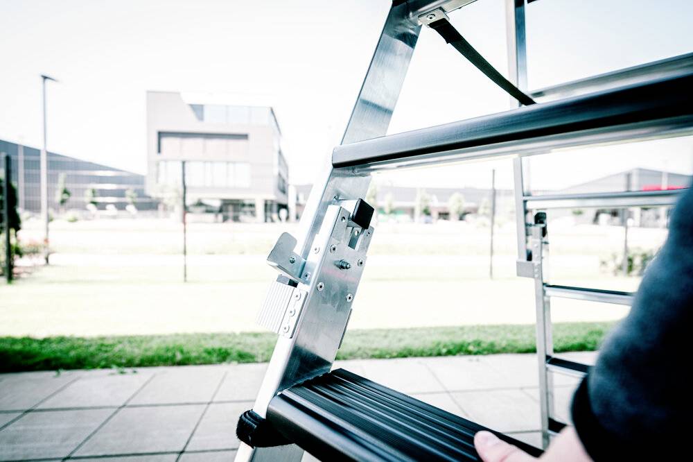 Osoba wspina się po srebrnej drabinie na wolnym powietrzu. W tle widać budynki i zieloną trawę.